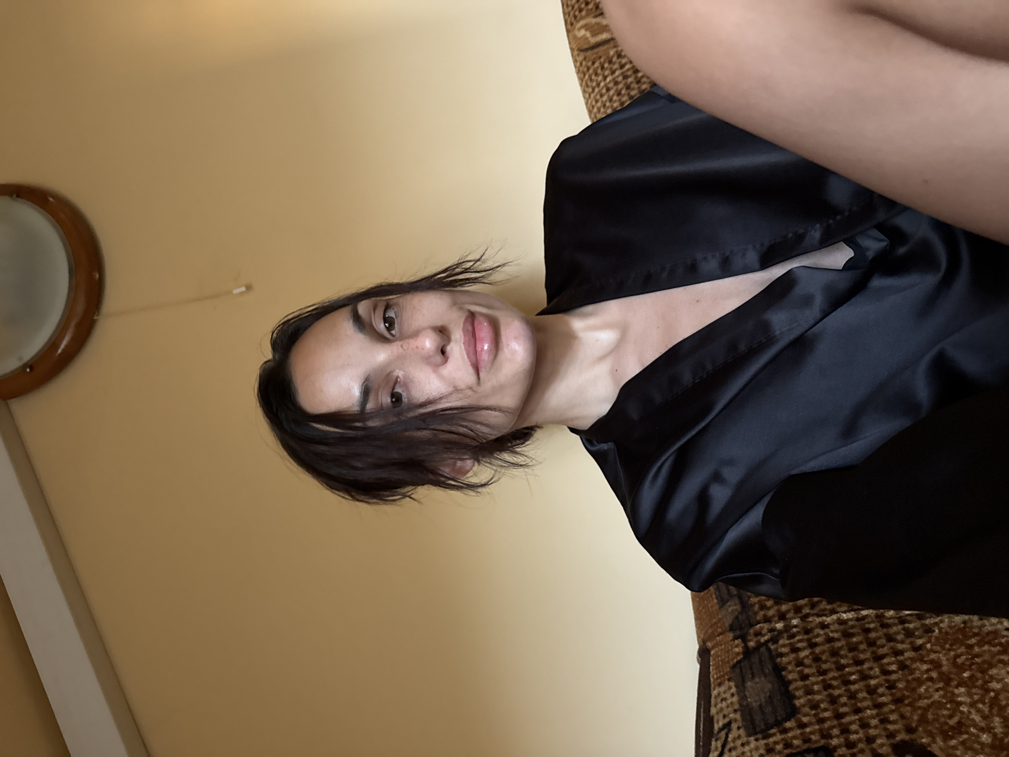 A woman with dark, messy hair, fair skin, and freckles is seen from the chest up, seated on a patterned sofa. She is wearing a black satin robe. The background consists of a yellow wall with a circular mirror mounted on it. The image appears to be taken indoors, in a residential setting. There is no visible indication of the city of Pleven, Bulgaria in the image.