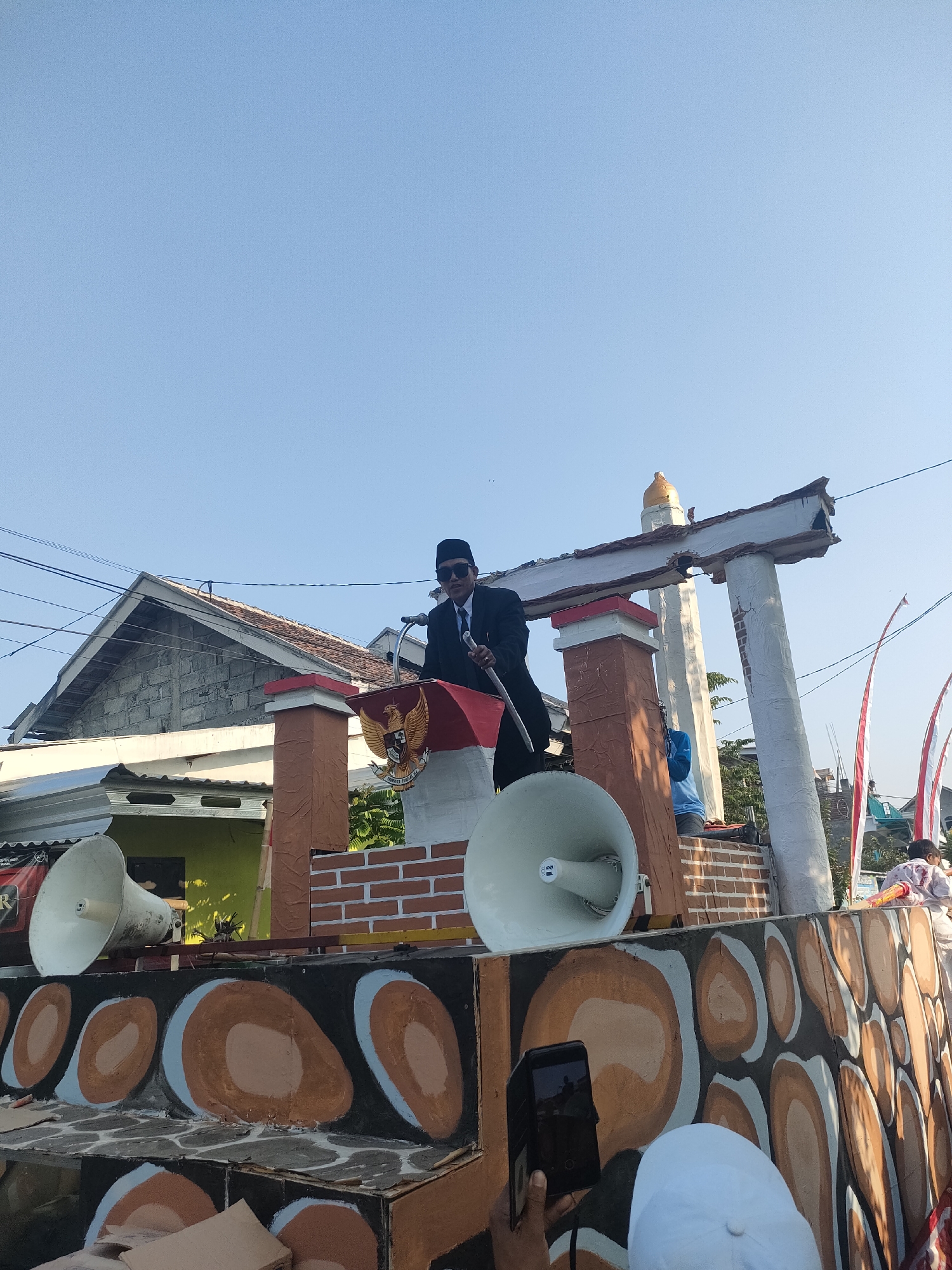 The image depicts an outdoor scene in Tongaswetan, Indonesia, likely during a public event or parade. In the foreground, a person dressed in a black suit, black hat, and sunglasses stands behind a podium adorned with the Indonesian coat of arms and a red and white flag. The individual is holding a baton.
The podium is part of a larger, decorated float or structure. Two large white loudspeakers are visible on either side of the podium. The float has a decorative base with painted circular and irregular shapes in brown and orange hues. In the background, residential buildings with tiled roofs and some overhead wires are visible.
Further in the background, a white structure resembling a gate with a golden dome on top stands. Red and white Indonesian flags are also seen planted in the background. Other people, including children, are present on the float and in the background, some of whom appear to be observing the scene. The sky is clear and blue, indicating a sunny day. The overall impression is of a festive or ceremonial gathering.
