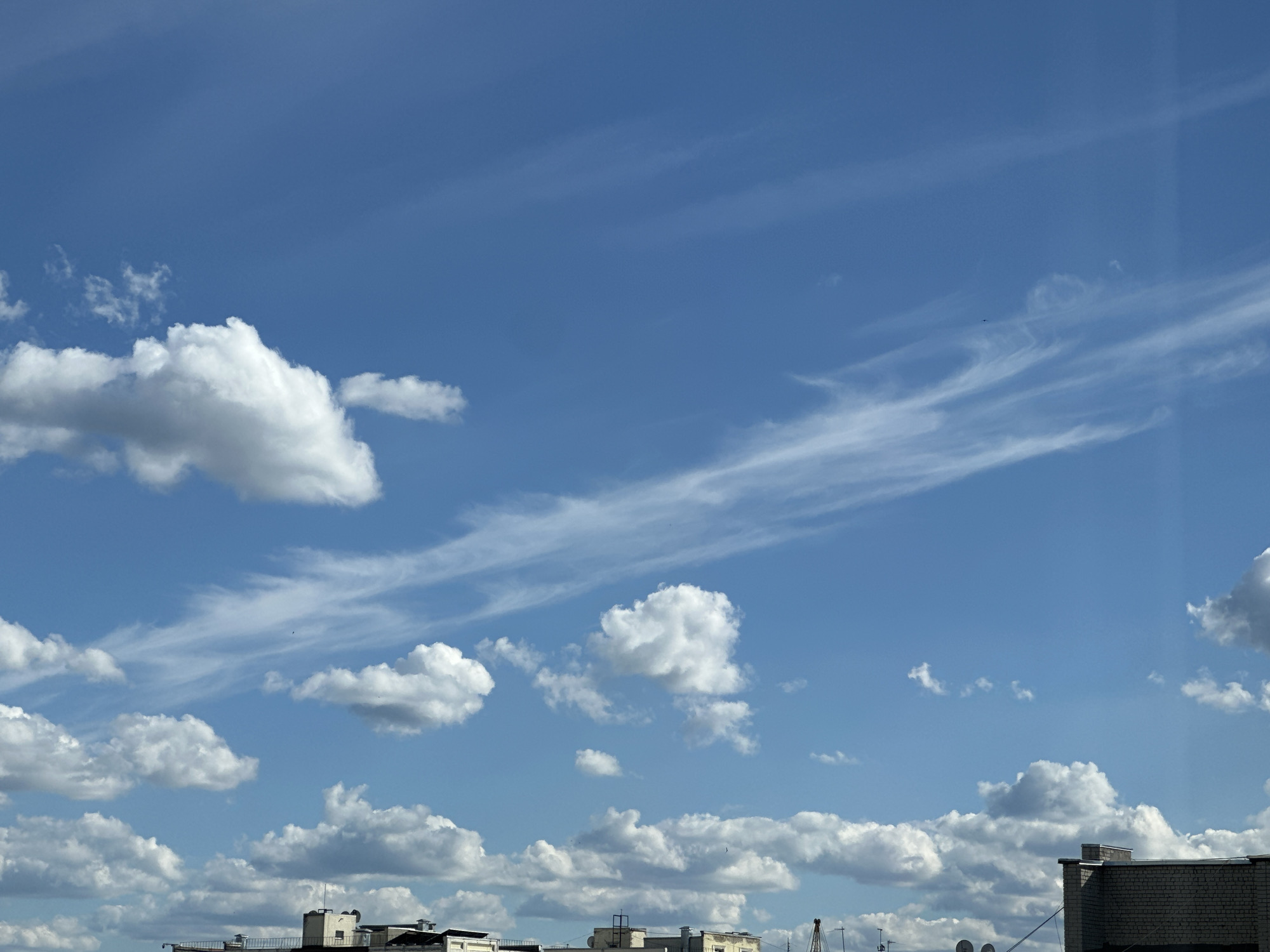 A wide-angle shot captures a bright blue sky filled with a variety of clouds. Scattered cumulus clouds, some appearing fluffy and white, others more wispy and elongated, drift across the frame. In the upper portion of the sky, thin, streaky cirrus clouds create a delicate, feathery pattern. The bottom of the image shows the rooftops of several buildings, with architectural details like chimneys and antennae visible. The lighting suggests a clear, sunny day.