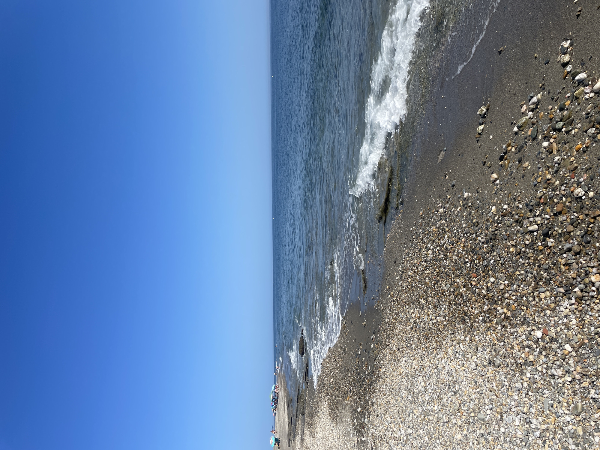 A wide-angle shot captures a serene coastal scene on a sunny day, likely on a beach in an unspecified location in Spain. The clear, deep blue sky dominates the upper half of the frame, meeting the calm, blue ocean at the horizon. The sea gently laps against a pebble and sand beach, with small waves creating white foam as they reach the shore.
The beach itself is composed of a mixture of small, smooth pebbles in various shades of grey, white, and brown, interspersed with darker sand. In the distance, to the left of the frame, several beach umbrellas and a few people can be seen, suggesting a sparsely populated recreational area. The water near the shore appears shallow, with some larger stones and darker patches visible on the seabed. The overall impression is one of tranquility and a typical summer beach day.