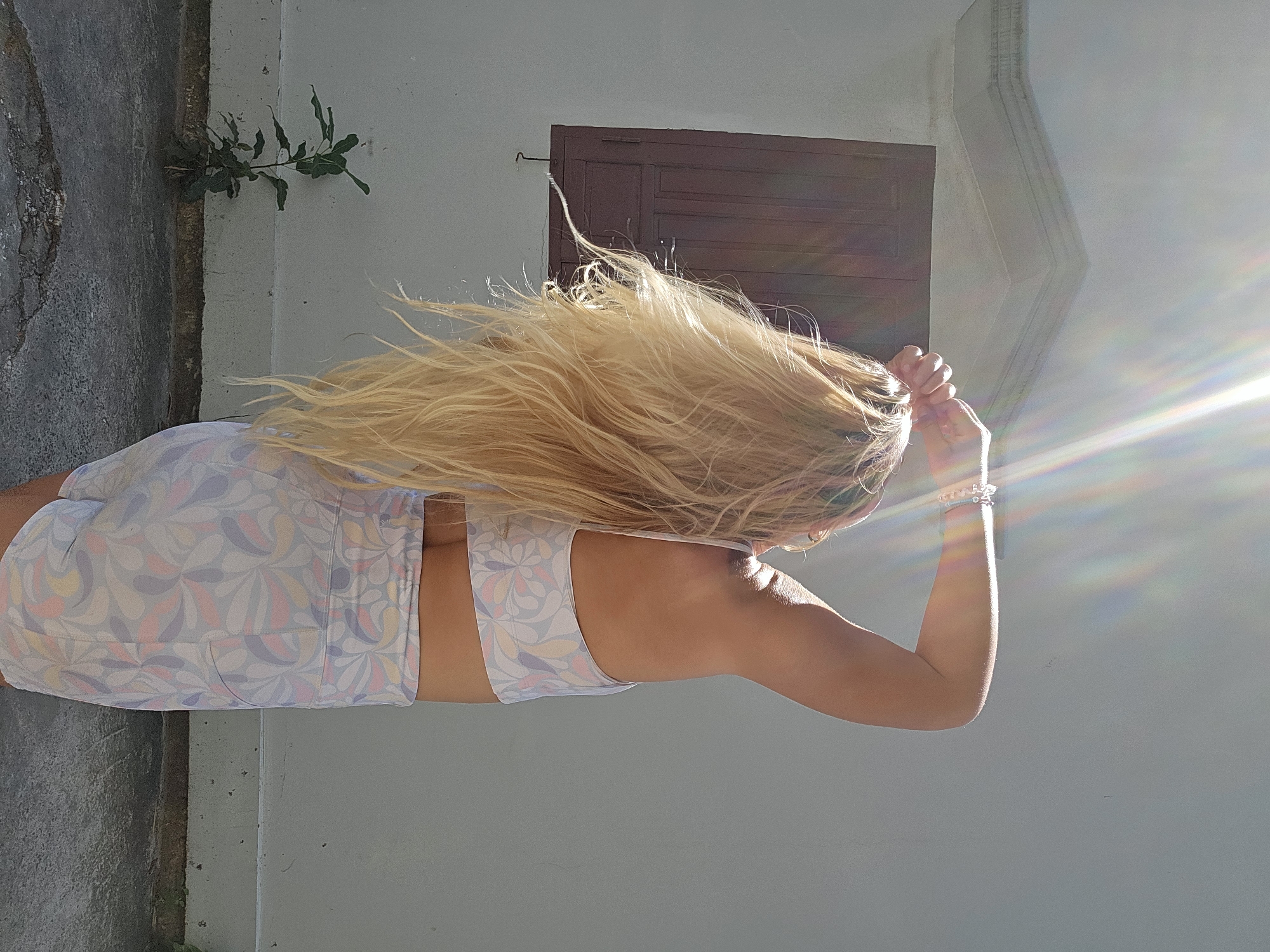 A person with long blonde hair, viewed from the back, is standing outdoors against a white wall. They are wearing a patterned sports bra and shorts. The person's right arm is raised behind their head, and their left hand is touching their hair. Sunlight streams down from the upper right, creating lens flare. A closed wooden window is visible on the wall behind the person. To the left, a small green plant grows from the ground in front of the wall. The ground appears to be cracked concrete. The scene suggests a bright, sunny day.
