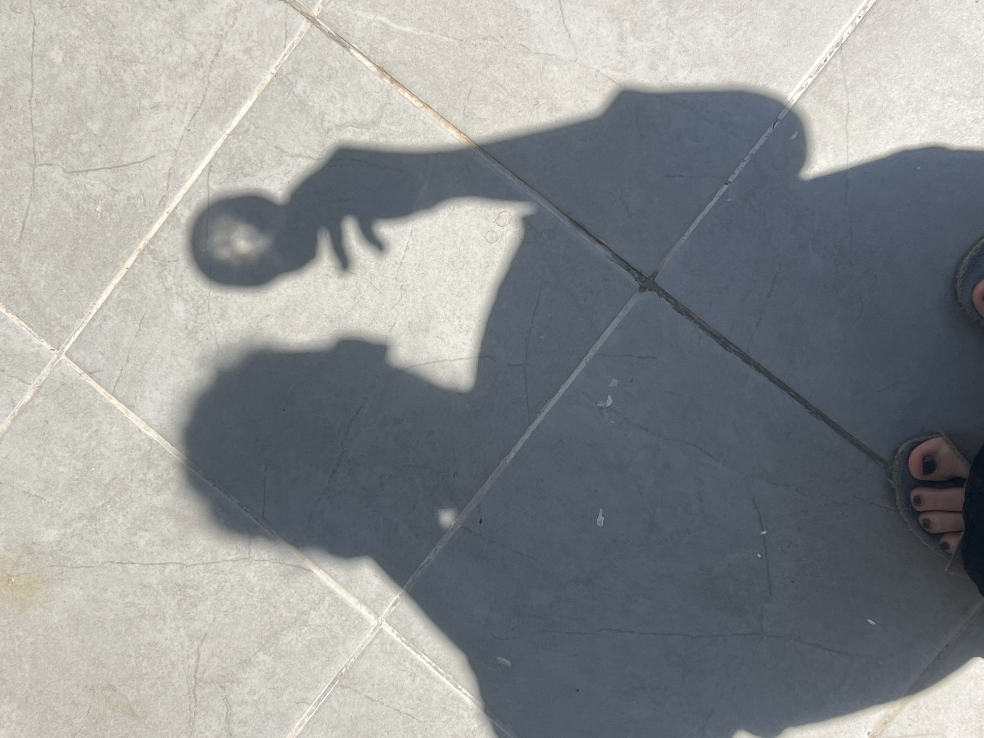 The image shows a shadow of a person on a tiled floor. The person appears to be holding a circular object, possibly a phone or a small accessory, in their right hand. The shadow is cast by direct sunlight, indicating an outdoor environment. The tiles are light gray and laid in a grid pattern. The bottom of the image shows the shadow of a foot wearing a sandal. There are no discernible landmarks or contextual clues to pinpoint a specific location within Tel-Aviv, Israel. The scene is static and devoid of any interaction or narrative beyond the implied presence of a single individual.