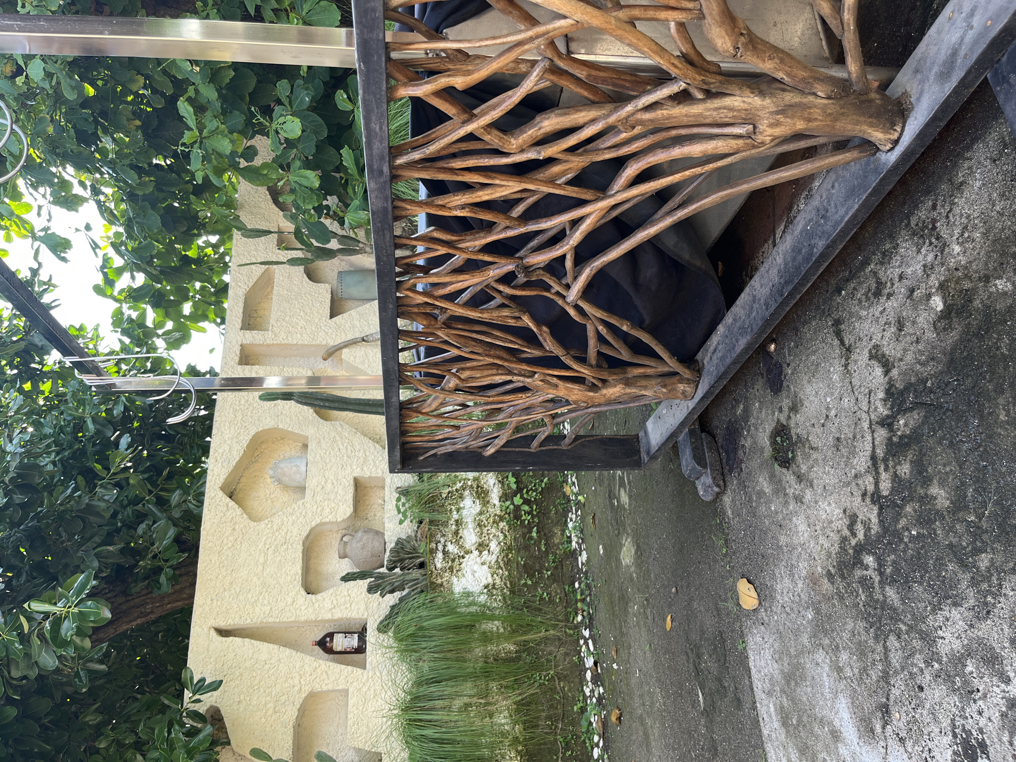 The image depicts an outdoor scene with a focus on a decorative wooden railing made of intertwined branches. Behind the railing, a portion of what appears to be a seating area is visible, covered with a dark fabric. To the left, a textured, pale yellow wall features several recessed niches. One niche holds a dark glass bottle with a label, while another contains an ancient-looking ceramic pot. Other decorative elements on the wall include some subtle sculptural shapes and what appear to be small cacti. In front of the wall, a lush layer of tall, green grass and various smaller plants creates a naturalistic border. The ground in the foreground is concrete, showing signs of wear and some scattered dry leaves. Above and behind the wall, dense green foliage from trees creates a shaded canopy. The overall impression is of an outdoor space, possibly a garden or a patio area in Kuta Selatan, Indonesia, characterized by a blend of natural and decorative elements.