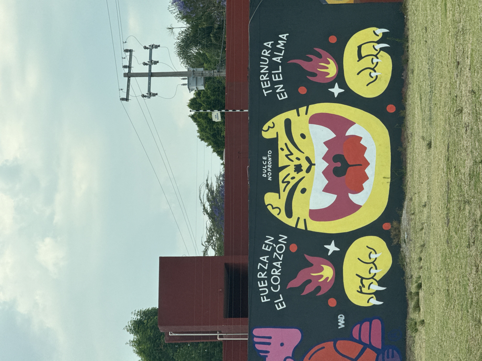A photograph taken in San Andrés Cholula, Mexico, shows a portion of a mural painted on a low wall adjacent to a grassy area. The mural, executed in a flat, graphic style, features a stylized tiger-like face in yellow with a crimson interior of the mouth, white teeth, and a black background. Above and below the face are yellow claws or paw prints with white claws, each accompanied by a small flame. The mural includes text in Spanish. "Ternura en el alma" (Tenderness in the soul) is above the tiger face, and "Fuerza en el corazón" (Strength in the heart) is below. The phrase "Dulce no pronto" is included near the center of the mural. The mural also has the signature "WAD" in the bottom left corner.
Behind the mural, a two-story maroon-colored building is visible. Above the building and mural is a utility pole with transformers and power lines. The sky above the utility pole is a pale blue with clouds. Green trees surround the building and the mural area.