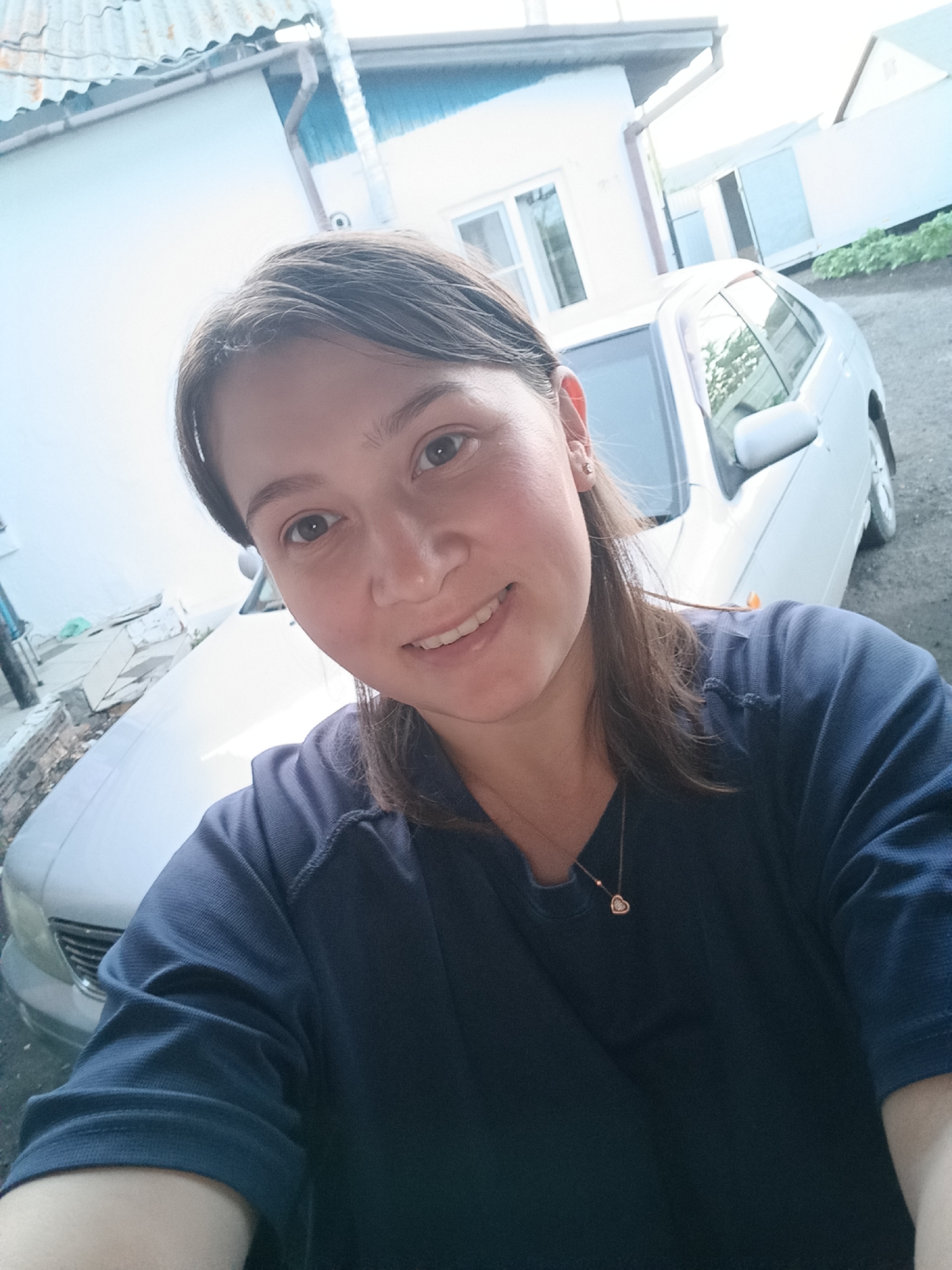 A woman is taking a selfie in front of a house and a car. She is wearing a dark blue shirt and a necklace with a heart-shaped pendant. The house appears to be a single-story building with a light blue and white exterior. A silver car is parked in the driveway. The scene suggests an outdoor setting, possibly a residential area. The location is specified as Хохлы, Russia.