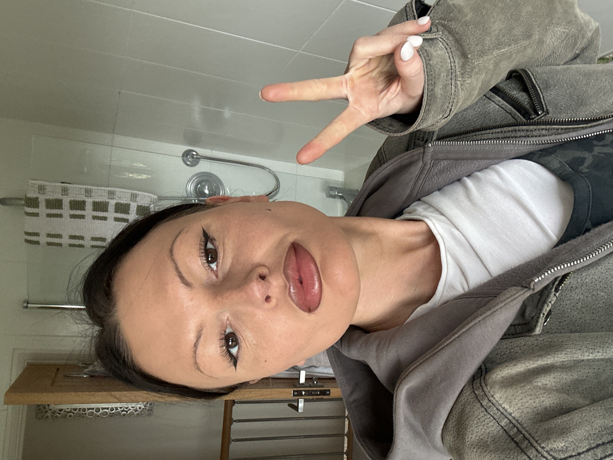 An adult female is depicted in a selfie, positioned indoors within the city of Wealden, United Kingdom. She has dark hair and brown eyes with winged eyeliner and defined eyebrows; her lips are glossy and slightly parted. She wears a white t-shirt, a grey hooded sweatshirt, and a grey-green textured zippered jacket, with a dark camouflage pattern visible beneath. Her left hand is raised, displaying a "peace" sign, with white-painted fingernails. The background on the right features light-colored wall panels or tiles, a shower head with a flexible hose, and a white and green patterned towel hanging on a chrome bar, indicating a bathroom. To the left, a partially open wooden door is visible, along with a decorative mirror featuring circular elements, and a metal railing.