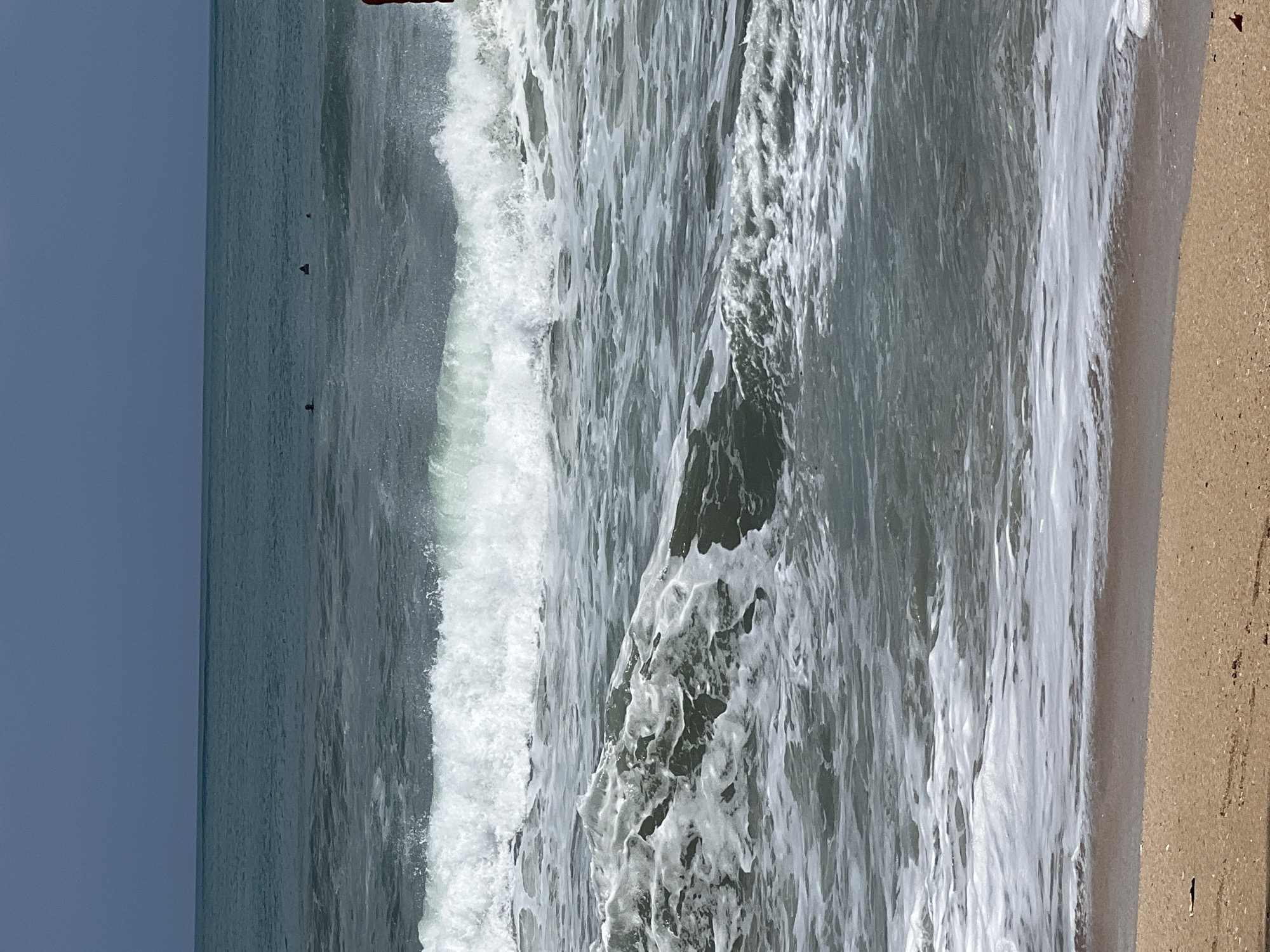 The image captures a dynamic ocean scene with breaking waves on a sandy beach. The water is a deep, slightly murky teal color, with white foam cresting the waves. In the distance, several small dark figures are visible in the water, suggesting people are swimming or surfing. The sky is a muted, overcast gray. The foreground shows a stretch of golden sand, with some small debris visible on its surface. The scene is devoid of any man-made structures or explicit landmarks that would indicate a specific location within Kuta, Indonesia, beyond it being a coastal area.