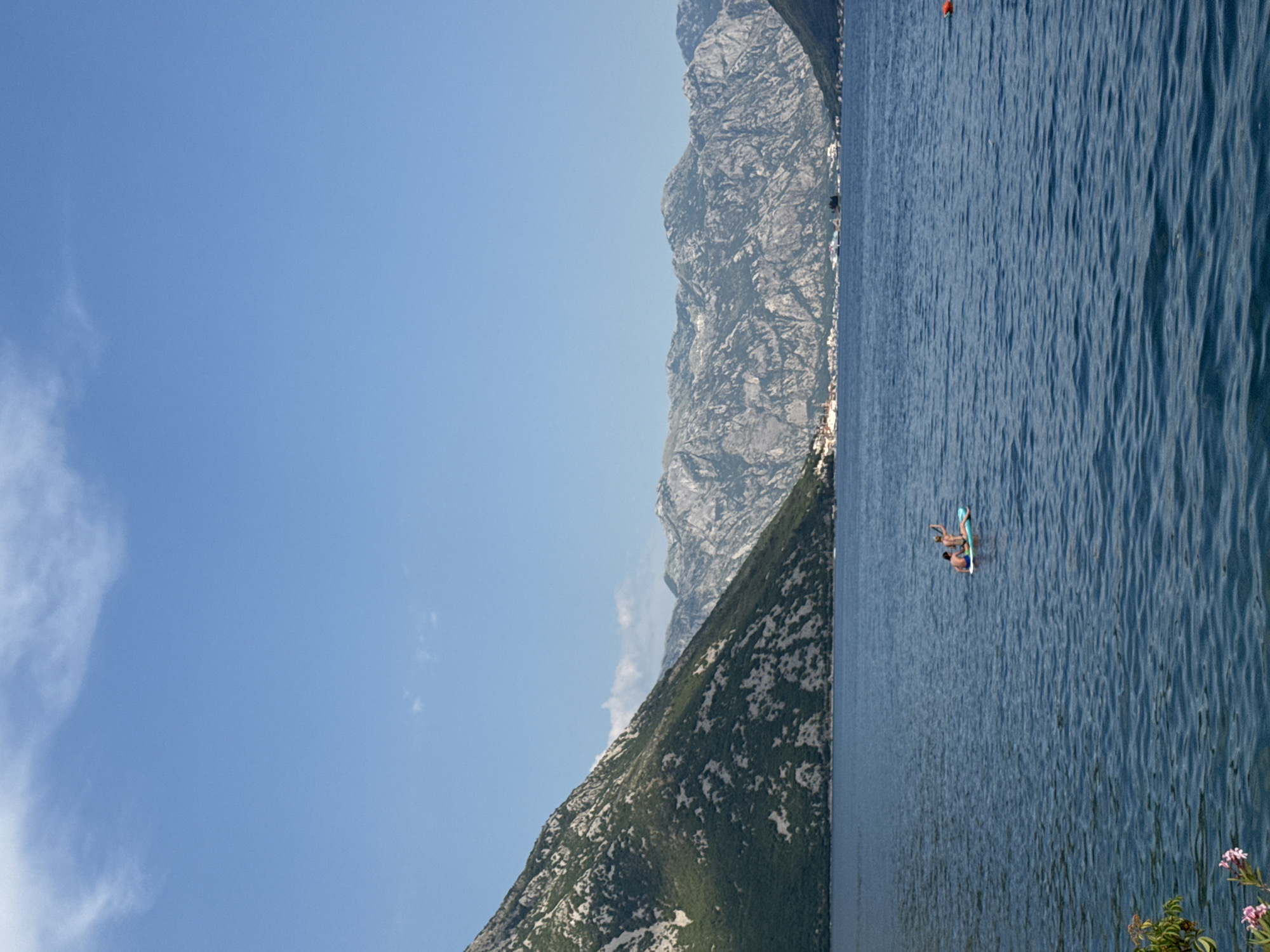 This is a landscape photograph taken in Lipci, Montenegro. The image depicts a vast body of calm, deep blue water on the right, with a steep, rocky mountain range covered in sparse greenery on the left. The sky above is a clear, bright blue with a few wispy white clouds on the far left. The sun appears to be high in the sky, suggesting it is midday.
In the water, two individuals are visible on a light-colored inflatable board. They appear to be enjoying a leisurely activity on the water. A single red buoy floats in the water in the upper right portion of the image.
The mountainous terrain dominates the left side of the frame, with sharp, textured rock formations. The water's surface exhibits gentle ripples, indicative of a calm day. In the lower right corner, a small cluster of pink flowers and green foliage adds a touch of natural detail to the scene. The overall mood is serene and picturesque, highlighting the natural beauty of the coastal mountainous region. There is no visible text in the image.