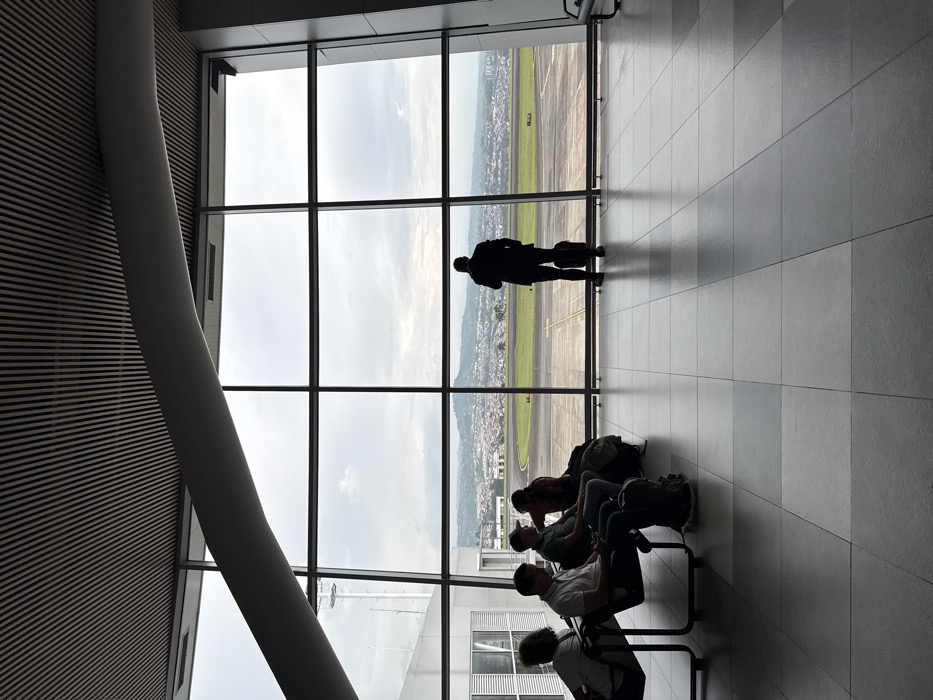 The image depicts the interior of a modern airport terminal waiting area at Matecaña International Airport (Pereira), located in the Area Metropolitana Centro Occidente (AMCO), Colombia. The scene is primarily illuminated by natural light entering through large, floor-to-ceiling windows that offer a panoramic view of an airfield and the distant city of Pereira.
The floor consists of alternating light and dark grey square tiles. Overhead, a slatted ceiling is visible, along with a prominent, curved, light grey structural beam traversing the upper portion of the frame.
On the left side, four individuals are seated in black, metal-framed airport waiting chairs. They are largely silhouetted against the bright exterior. From left to right: an individual with lighter hair is partially visible; a male in a light polo shirt looks downwards, with a dark backpack positioned on the floor in front of him; a male wearing a baseball cap and a dark t-shirt looks towards the right; and another individual, possibly female, with long hair, is seated further back, with a large bag on the floor next to them.
On the right side, a solitary male figure stands facing away from the viewer, looking out the windows. He is dressed in a dark suit jacket and trousers, and a dark, wheeled suitcase rests at his feet to his right.
Through the windows, a section of an airfield runway or taxiway with faint markings is visible in the mid-ground. Beyond the airfield, the urban landscape of Pereira extends across rolling hills, reaching towards a distant mountain range. A white tower structure with antennae is visible through the windows on the far left. The sky is overcast with a consistent grey cloud cover, indicating daytime under cloudy conditions.