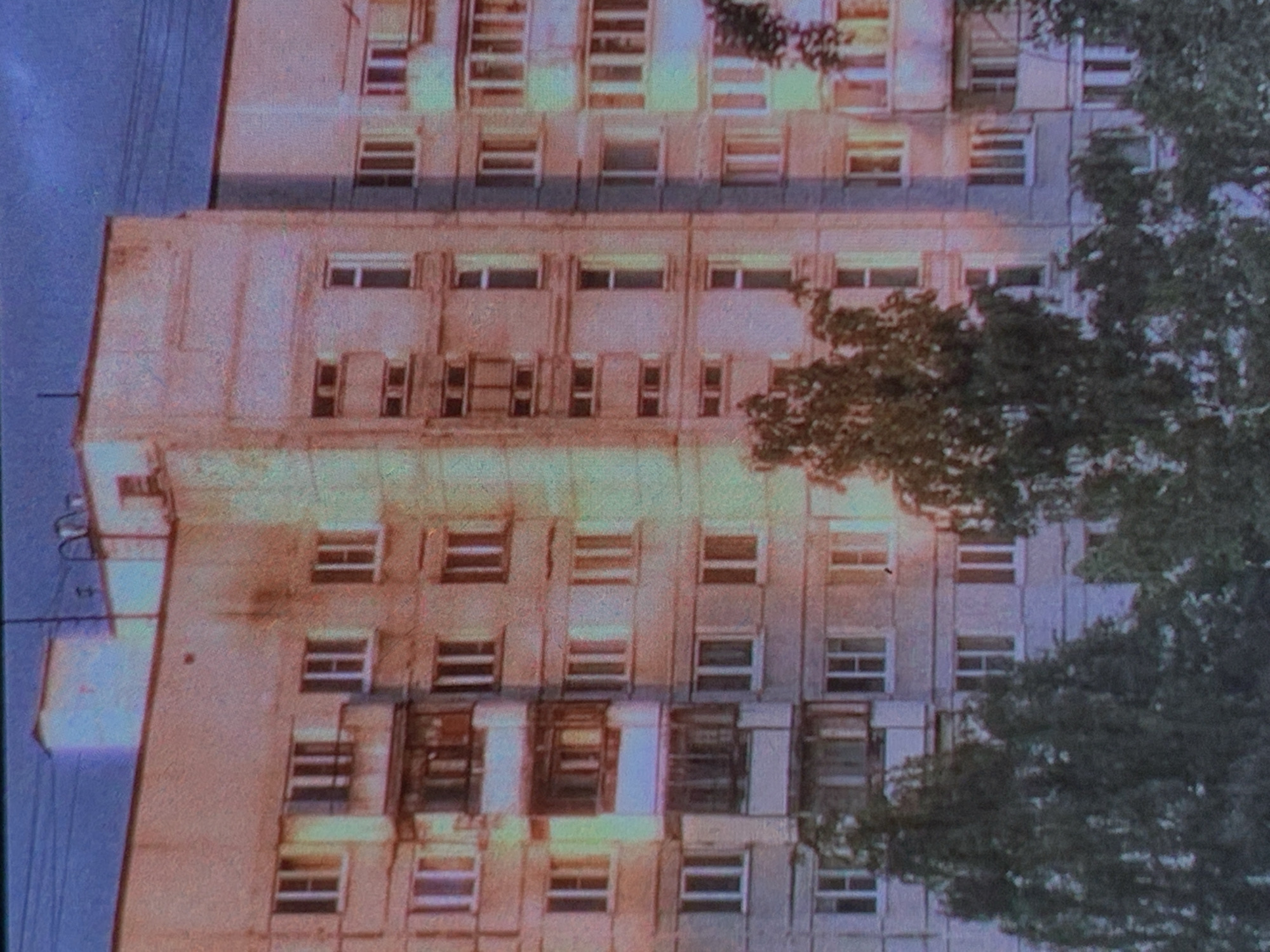 The image shows the facade of a multi-story residential building in Nizhny Tagil, Russia, with trees in the foreground. The building appears to be constructed from concrete panels and has a repetitive pattern of windows and balconies. The overall lighting suggests it is either early morning or late evening, with a purplish hue in the sky. There are no people or discernible activities visible in the image. The style of the building suggests it is a typical Soviet-era apartment block.
