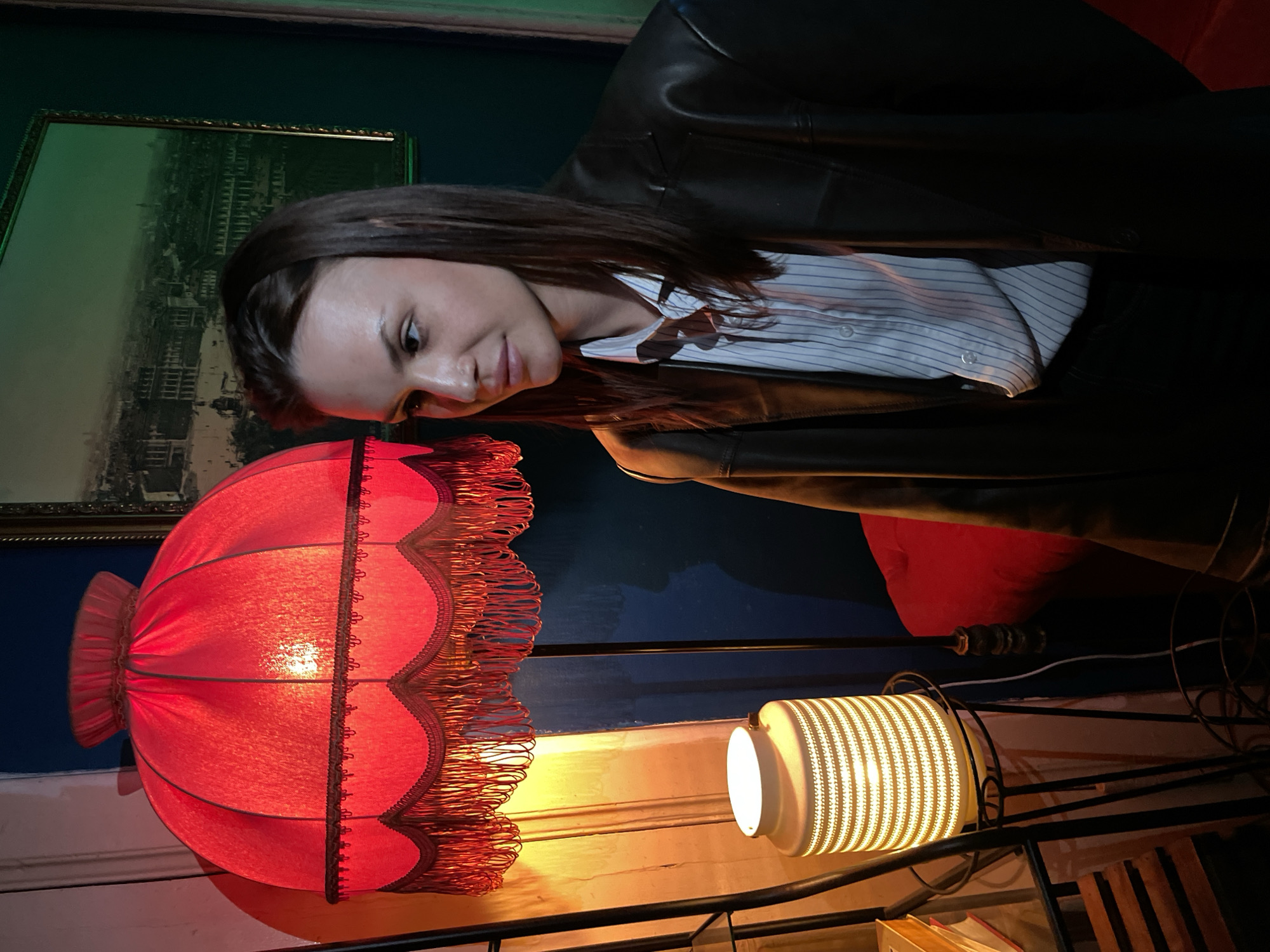 The image depicts a woman indoors, likely in Saint Petersburg, Russia, based on the style of the setting. She is wearing a black leather jacket over a striped shirt. A red fringed lampshade dominates the foreground, with a smaller white cylindrical lamp also visible. A framed picture hangs on the wall in the background. The scene is dimly lit, with both lamps providing the main sources of illumination.