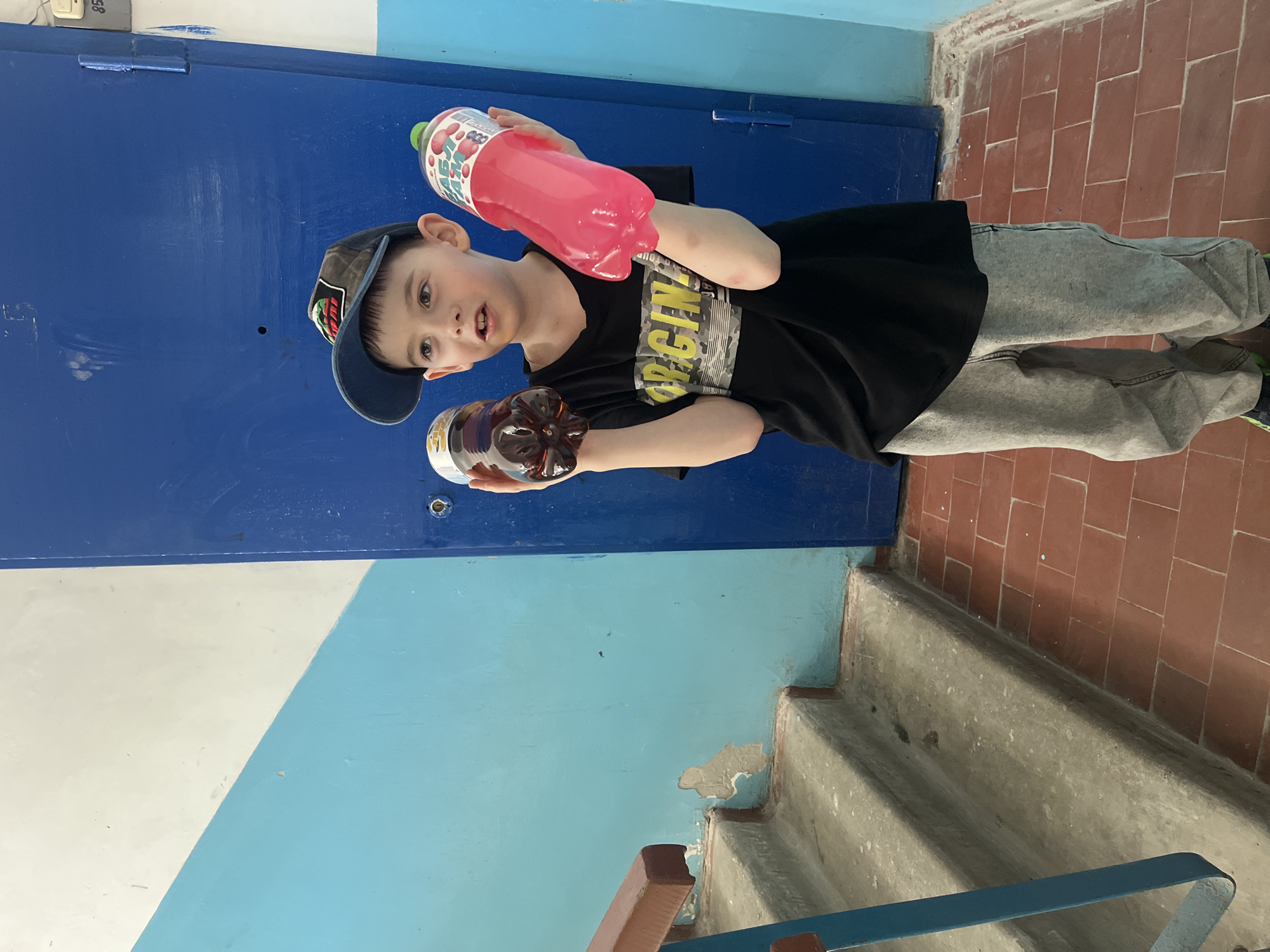 A young boy wearing a black t-shirt, grey sweatpants, a baseball cap, and green sneakers is holding a pink bottle in his right hand and a brown bottle in his left hand. He is standing on a set of stairs in a building in Chaikovsky, Russia. The wall behind him is painted blue and white, and the door behind him is painted dark blue. The stairs are tiled with brown tiles.
