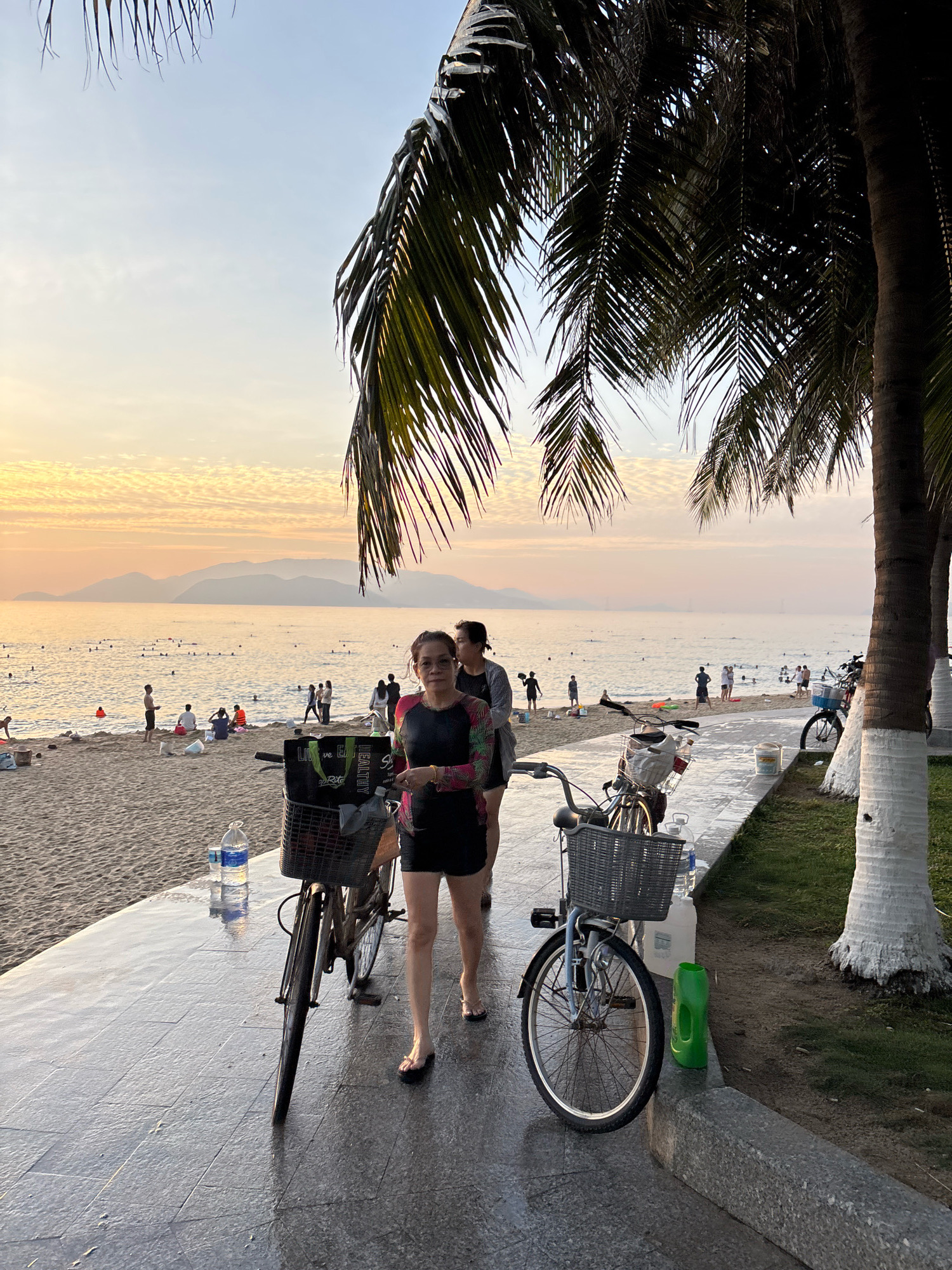 The image depicts a beachfront scene in Nha Trang, Vietnam, during a sunset. In the foreground, two bicycles are parked on a paved walkway adjacent to a sandy beach. A woman wearing a multi-colored long-sleeved shirt and black shorts stands between the two bicycles, looking towards the camera. Behind her, another person is partially visible. The beach is populated with numerous individuals, some swimming in the ocean and others relaxing on the sand. The ocean is calm, and mountains are visible in the distance, silhouetted against the colorful sunset sky. Palm trees frame the top and right side of the image. Several bottles of water and a green container are placed near the bicycles. The overall atmosphere suggests a relaxed evening at the beach.