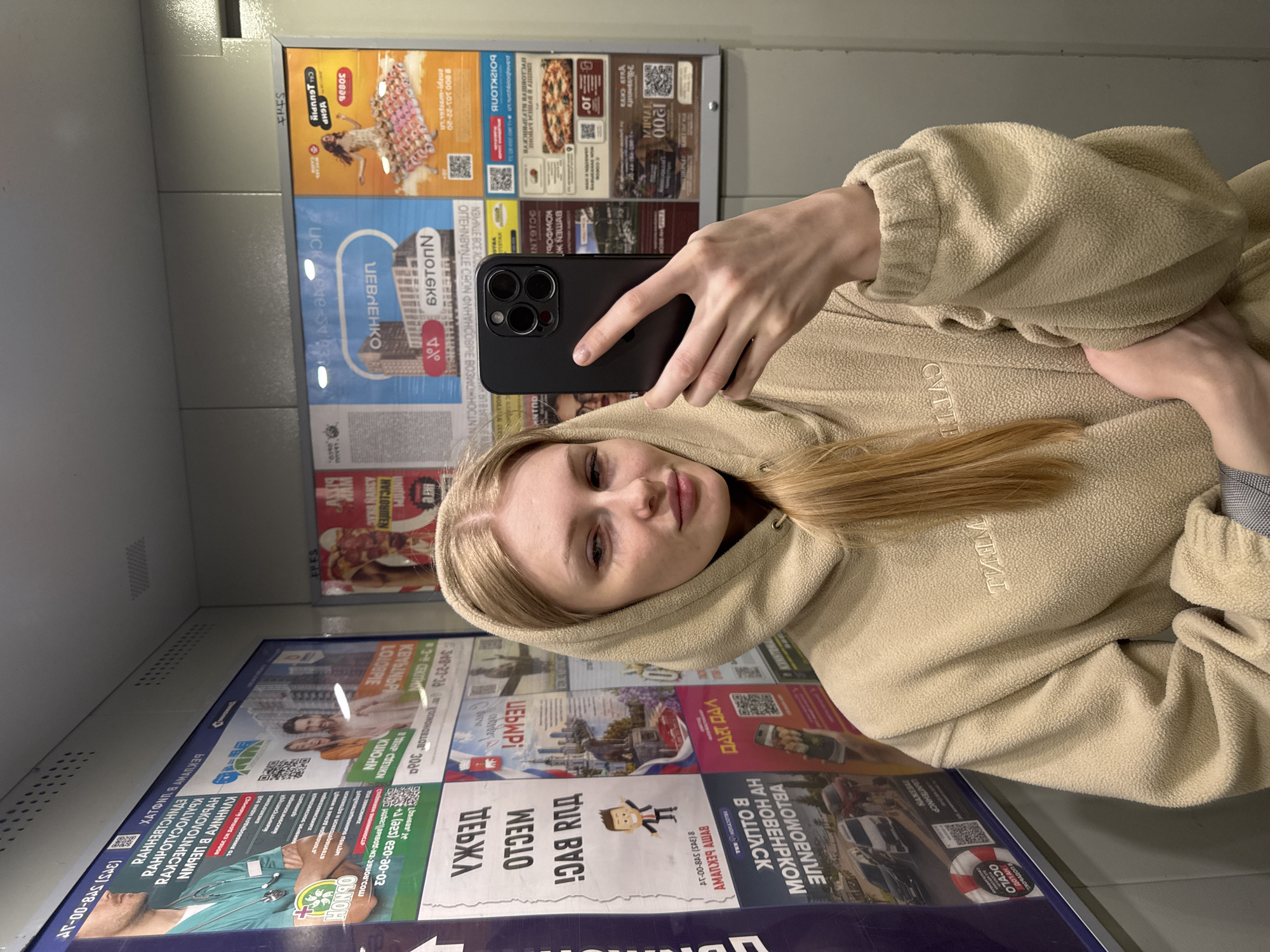 In a city elevator in Kondratovo, Russia, a young woman with blonde hair is taking a selfie. She is wearing a light beige hooded sweatshirt, the hood of which is pulled up over her head. Her right hand holds a black smartphone, angled to capture her reflection in the elevator's mirrored walls. The elevator walls are adorned with various advertisements, including posters for medical services, new housing developments, and food establishments like pizza and sushi. The lighting is dim, typical of an elevator, casting a subtle shadow on the woman's face. The overall scene is an everyday moment captured within the confined space of a public elevator.
