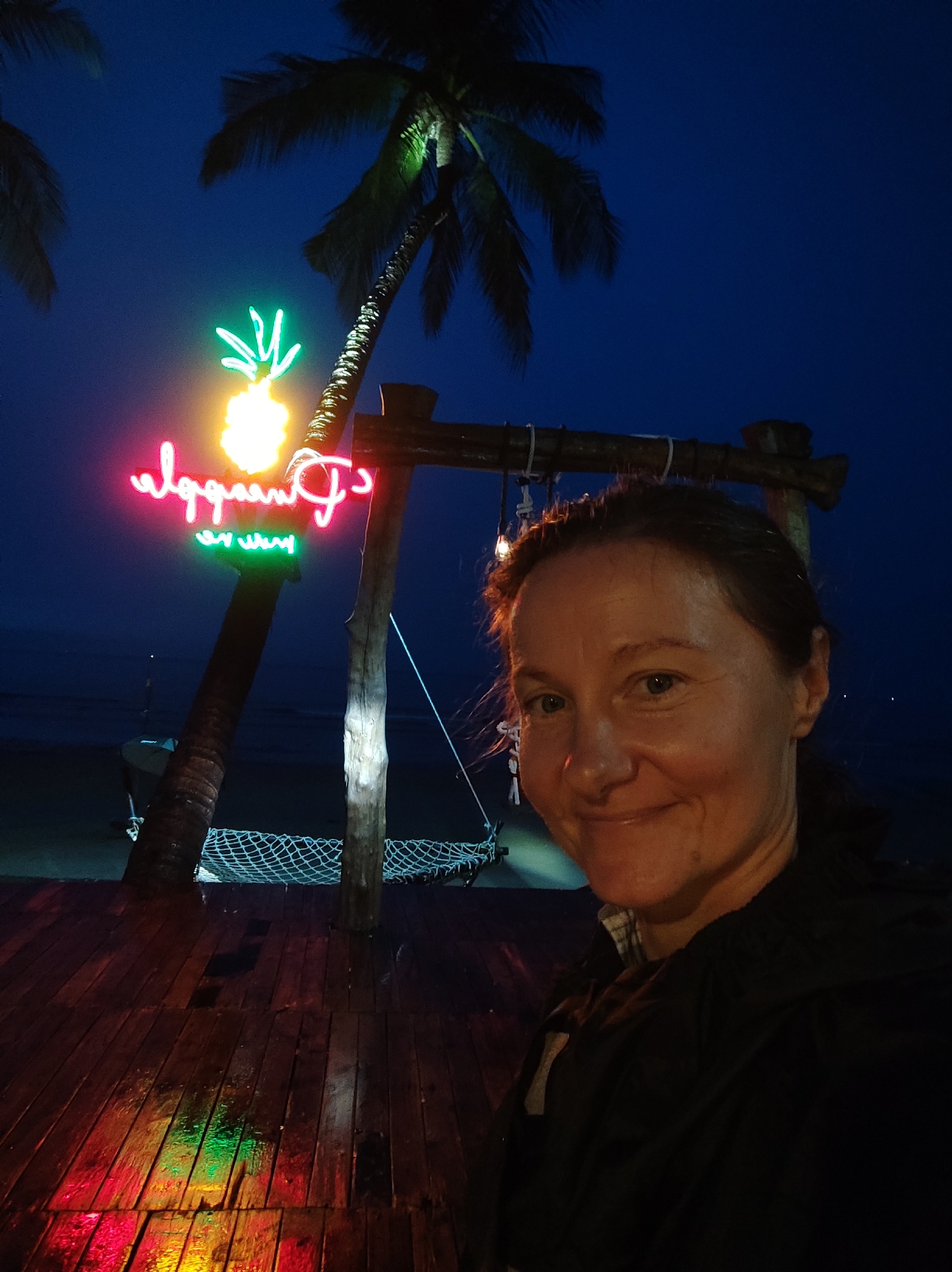 A medium shot captures a woman at night on a wooden deck, likely a seaside bar or restaurant in Unknown, Unknown. Her face is partially lit, revealing fair skin, blue eyes, and brown hair pulled back. She's wearing a dark jacket, and part of a light-colored shirt is visible beneath it.
Behind her, a palm tree stands tall, supporting a brightly lit neon sign shaped like a pineapple with lettering beside it. The reflection of these lights illuminates the wet wooden deck. A hammock is suspended behind the woman. The backdrop includes the ocean, barely visible under the dark sky.