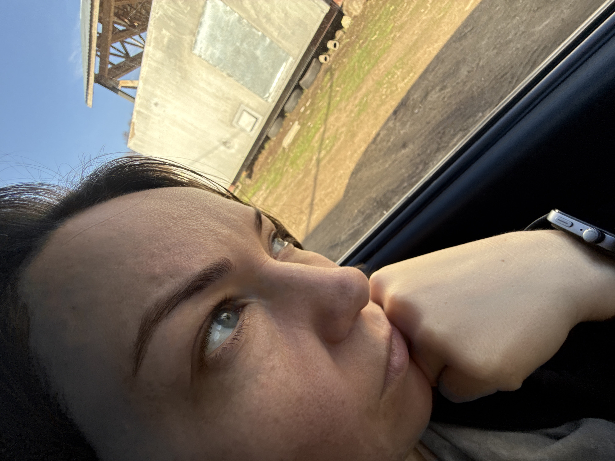 The image presents a heavily tilted, close-up perspective of a person looking out of a vehicle window on a clear day in Chaikovsky, Russia. The main subject is an adult female with dark hair and light blue eyes, whose face fills much of the foreground, positioned diagonally across the frame. She gazes upwards and outwards with a pensive or contemplative expression, her right hand balled into a fist and resting against her mouth or chin. A silver-colored smart watch, likely an Apple Watch, is visible on her left wrist.
Through the vehicle window, a bright blue sky is prominent on the left side of the frame. The background outside the vehicle features a light-colored, weathered building, possibly concrete or metal, with a reflective rectangular panel. Above and slightly behind this building, a wooden structural element or roof truss is visible against the sky. Below the building and extending to the right, an expanse of light brown ground with patches of green grass stretches towards what appears to be a dark, paved road or path. Several small, cylindrical objects, possibly pipes or pots, are seen near the building's base. The lighting suggests it is daytime, possibly late morning or afternoon, with clear weather. The overall scene conveys a moment of quiet reflection during travel. No visible text is discernible in the image.