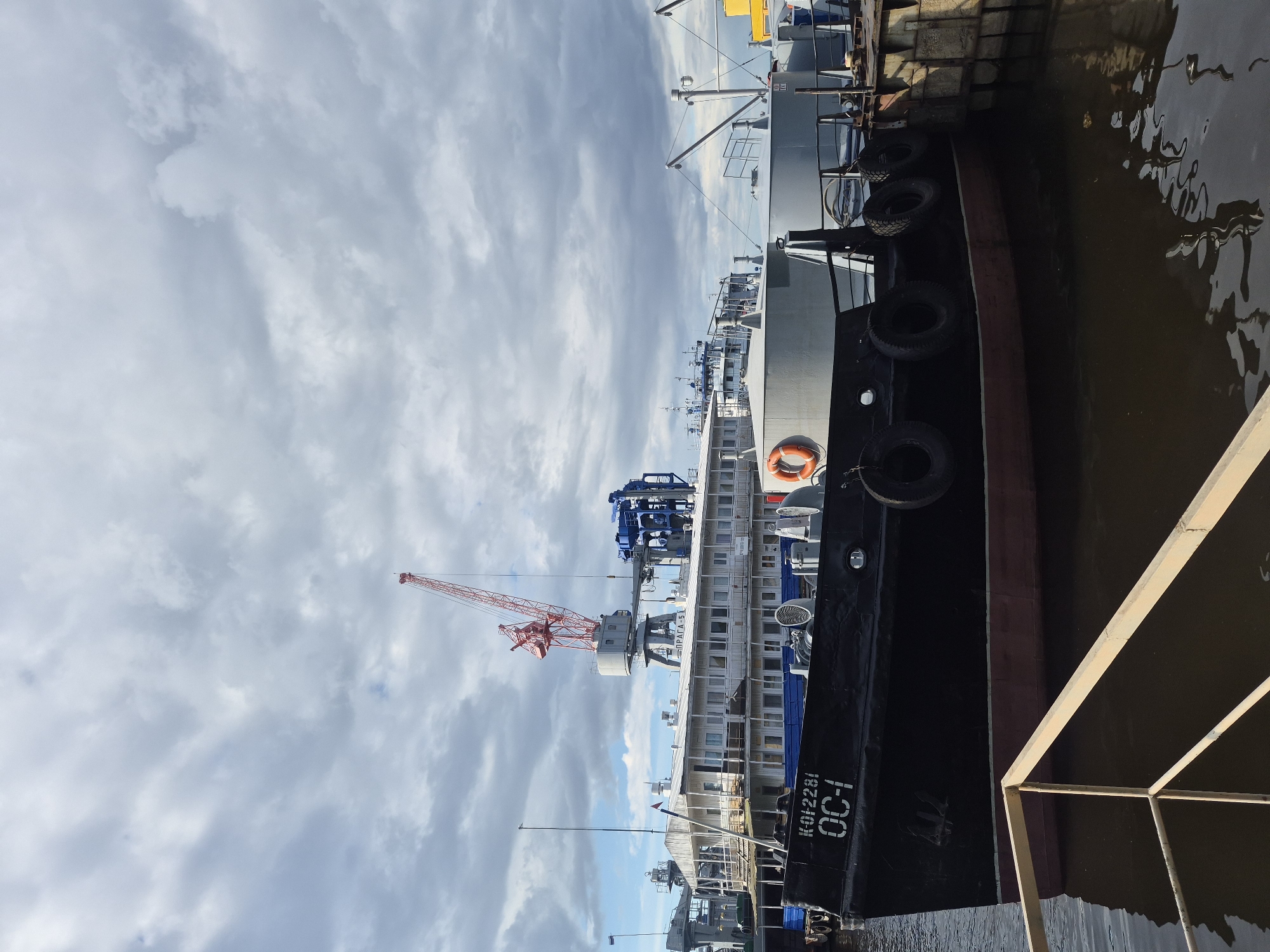 The image depicts a view of ships docked in what appears to be a harbor or port area, with a cloudy sky overhead. In the background, one can see construction cranes and other shipyard equipment. The location is likely Chaikovsky, Russia. A vessel with the designation "OC-1" and "K012281" is docked and visible. There is also a portion of a crane labeled "ПРAГA-5". The water surface reflects the sky and surroundings. The perspective is tilted, giving an unusual view of the scene.