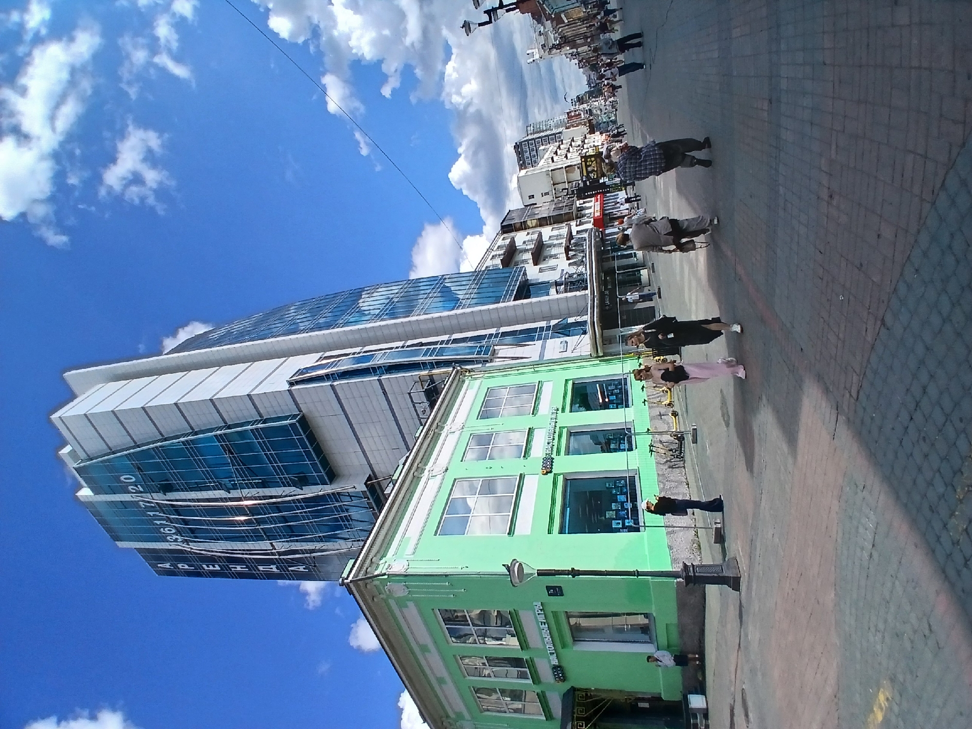 This media file captures a bustling street scene in Yekaterinburg, Russia, under a bright, cloudy sky, suggesting it's daytime. The perspective is low and tilted, giving a dynamic feel to the urban environment.
The foreground features a vibrant, light green building with multiple windows, likely a commercial establishment given the signage visible. Text on this building includes Cyrillic letters that appear to read "НЕ ТОЛЬКО ИГРЫ" ("NOT ONLY GAMES"). A lamppost stands near the building.
Dominating the left side of the image is a modern, multi-story building with a striking facade of blue glass panels and white architectural elements. Text is visible on this building, including "АРЕНДА" ("RENT") and a phone number.
The street is paved with cobblestones, and a crowd of people is walking along the sidewalk. They are diverse in age and attire, some appearing to be engaged in conversation or shopping. A few individuals are closer to the camera, providing a clearer view of their clothing and stances. One person in the foreground wears a dark outfit, while another wears a pink skirt and a dark top.
In the background, more buildings of varying architectural styles line the street, extending into the distance. The presence of power lines and streetlights indicates an urban infrastructure. The overall atmosphere seems lively and active, typical of a public street on a pleasant day. The clear blue sky with scattered white clouds suggests fair weather.