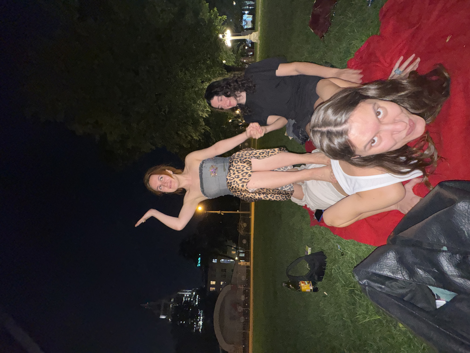 The image shows three individuals lying on a red blanket in a grassy area at night. One individual is wearing a white tank top, and another is wearing a grey top and a leopard-print skirt. The third individual is wearing a black t-shirt. They are holding hands. There are bags, bottles, and other objects scattered on the grass. In the background, there are buildings and streetlights, potentially indicating a city park or urban environment. The location is in Moscow, Russia.