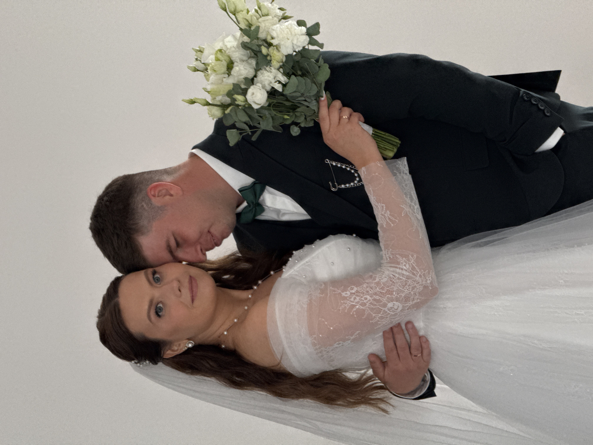 The image captures a newly married couple, a bride and groom, posed in a tender embrace, likely during a wedding ceremony or photoshoot in Chaikovsky, Russia. The photograph is oriented vertically, appearing as if the subjects are lying down against a plain, light background, though they are likely standing or seated with the camera tilted.
The groom, on the left, has short, dark hair and is dressed in a dark suit with a white shirt and a dark green bowtie. He leans his head towards the bride, seemingly kissing her forehead or whispering to her, with a gentle expression. A bouquet of white flowers, including roses and lisianthus, interspersed with green eucalyptus foliage, is held by his left hand, which rests on the bride's arm. A decorative pin with a chain and pearls is visible on his lapel. He wears a wedding band on his left ring finger.
The bride, positioned slightly below and in front of the groom, has long, dark brown hair that flows over her shoulder. She is wearing a white wedding gown featuring intricate lace on the long sleeves and a bodice adorned with sheer fabric and small pearls. A delicate pearl necklace and pearl drop earrings complement her attire, and a wedding veil is visible behind her head. Her left hand, also bearing a wedding band, rests gently on the groom's arm. She looks towards the camera with a serene and slightly smiling expression, her blue eyes open.
The background is a simple, uniform light grey or white, providing a clean and uncluttered setting that keeps the focus entirely on the couple. The overall mood of the image is intimate, affectionate, and joyful. There is no visible text in the image.