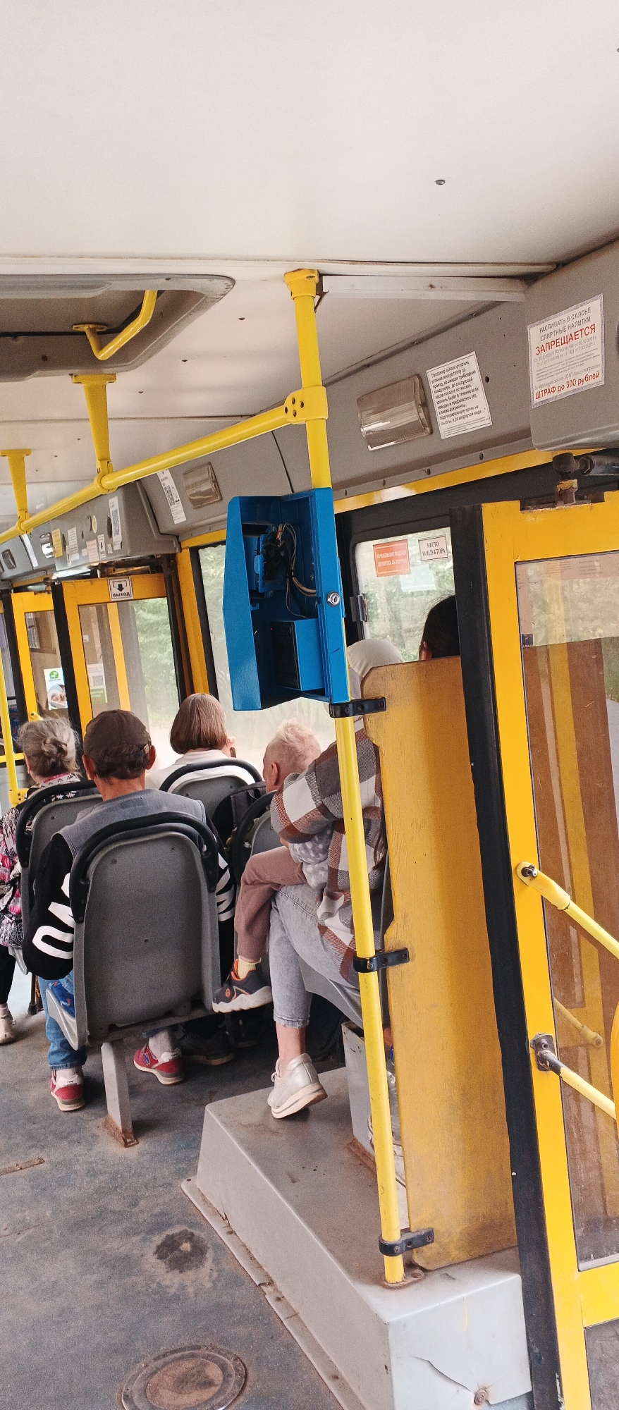 The image shows the interior of a public bus in Izhevsk, Russia. Several passengers are seated and standing. In the foreground, the worn floor of the bus is visible, with some dirt and a circular metal cover. Yellow handrails and support poles are prominent. A blue metal box, likely containing a ticket validator or other equipment, is mounted on a yellow pole. The seats appear to be made of dark grey plastic.
In the middle ground, passengers are seen from behind. A man wearing a grey vest over a black and white striped shirt and jeans is seated, facing away from the camera. A young child is being held on the lap of a person seated to the right, who is wearing jeans and a plaid shirt. Other passengers are visible further back.
In the background, through the windows, trees and foliage can be seen, indicating the bus is likely in motion or parked near a green area. A sign with Russian text is visible on the upper right side of the bus, and another sign with a pictogram and Russian text is on the left side near the doors. The overall impression is of a typical scene inside a city bus.