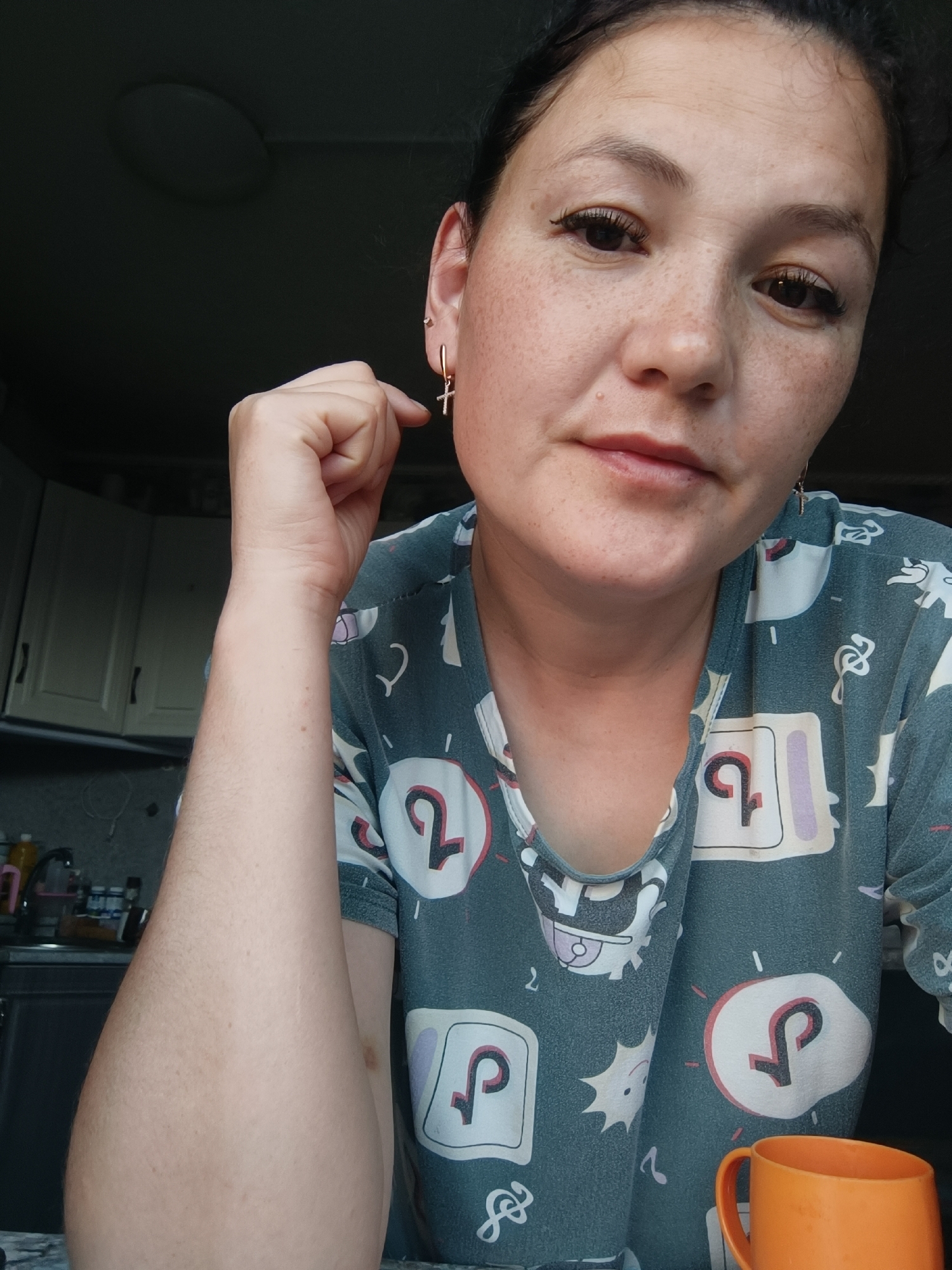 A medium shot shows a woman from the chest up, looking towards the camera. She has dark hair pulled back and is wearing a grey t-shirt with a repetitive pattern of white and pink cartoonish graphics, some resembling the TikTok logo. She has fair skin with freckles across her nose and cheeks and is wearing a small gold cross earring in her left ear. Her right arm is bent at the elbow, with her hand near her face, and her left arm is visible along the side of the frame, resting on a surface that is out of focus. In the lower right corner of the frame, a small part of an orange mug is visible. The background is dark and appears to be an indoor setting, with hints of kitchen cabinets and a ceiling light fixture. The image is likely taken in a residential kitchen.