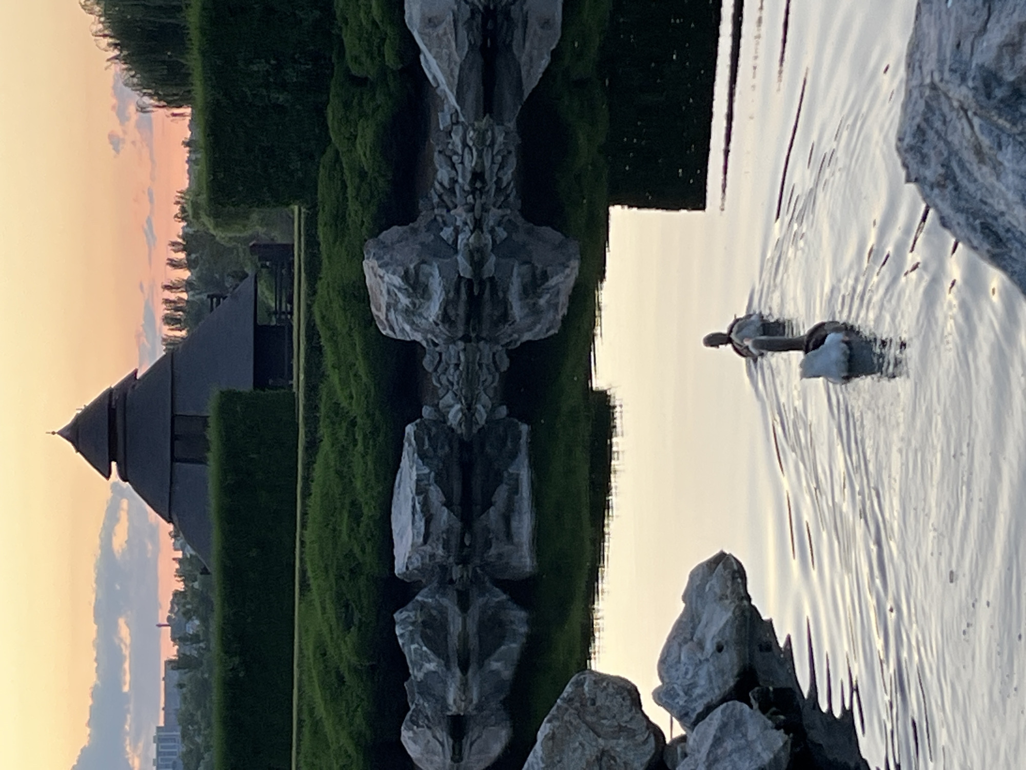 The image captures a serene park scene at dusk, likely located in the city of Dnipro, Ukraine, given the architectural style of the thatched-roof building and the surrounding flora. In the foreground, two ducks are swimming in a calm body of water, leaving gentle ripples in their wake. The water reflects the muted colors of the sky and the surrounding greenery. Large, grey rocks are strategically placed along the edge of the water, some submerged and others forming part of the bank.
In the midground, a meticulously trimmed green hedge forms a backdrop to the water. Behind the hedge, a distinctive building with a conical, thatched roof stands out, reminiscent of traditional architectural designs. The setting sun casts a warm, soft glow across the sky, with subtle hues of orange, pink, and pale blue visible through scattered clouds. The silhouette of trees and distant urban buildings can be seen on the horizon, suggesting the park is located on the outskirts of a city. The overall atmosphere is one of tranquility and natural beauty, enhanced by the soft light of the fading day.