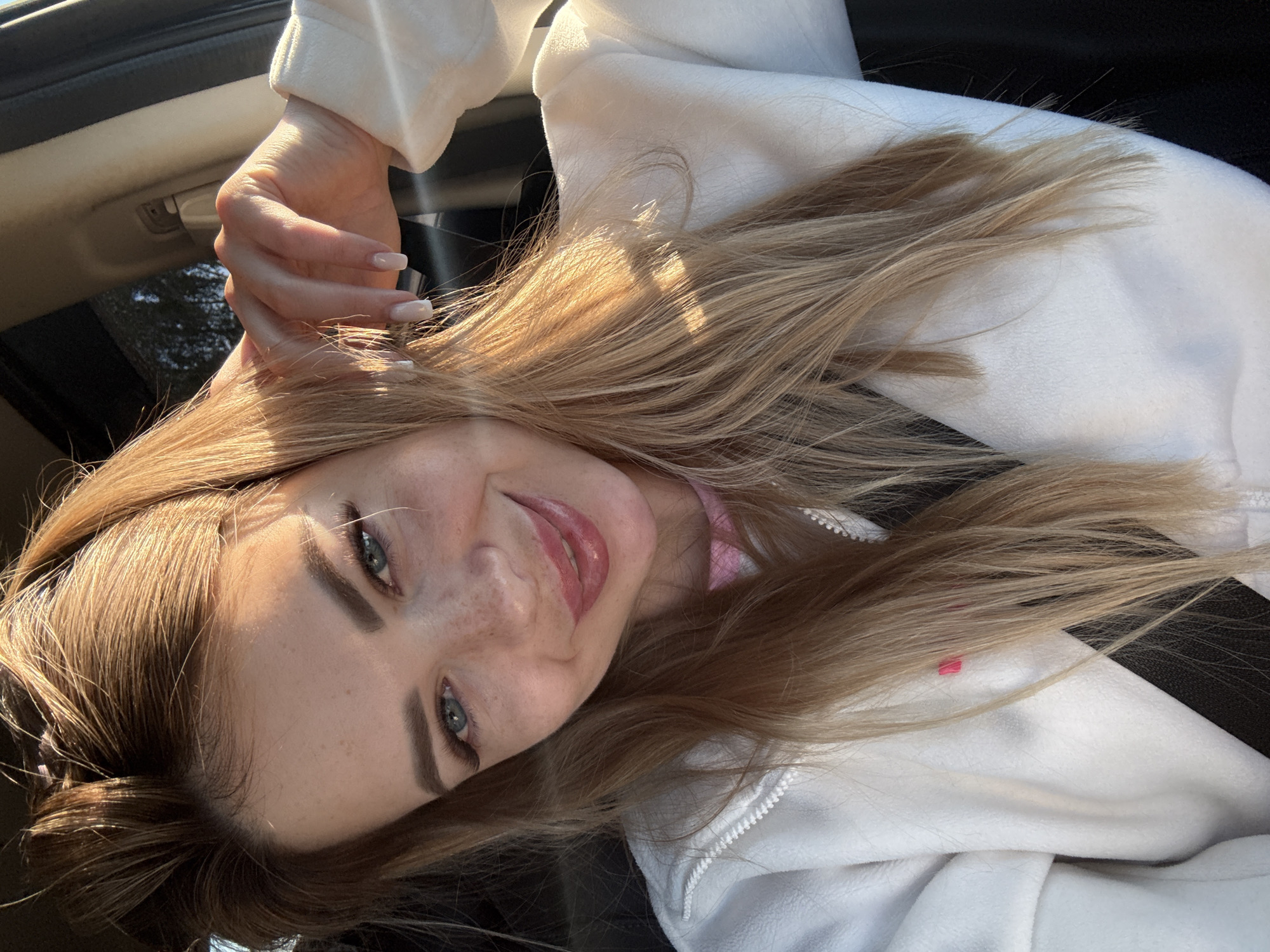 A young woman with blonde hair and blue eyes is sitting in a car, possibly in the passenger seat. She is smiling and looking at the camera, with her right hand near her face. She is wearing a white hooded jacket with a pink undershirt visible at the neckline. The sun is shining on her face and hair, creating a warm glow. The background is blurred and appears to be the interior of a car with a window showing trees outside. There are no other people or significant objects in the frame.