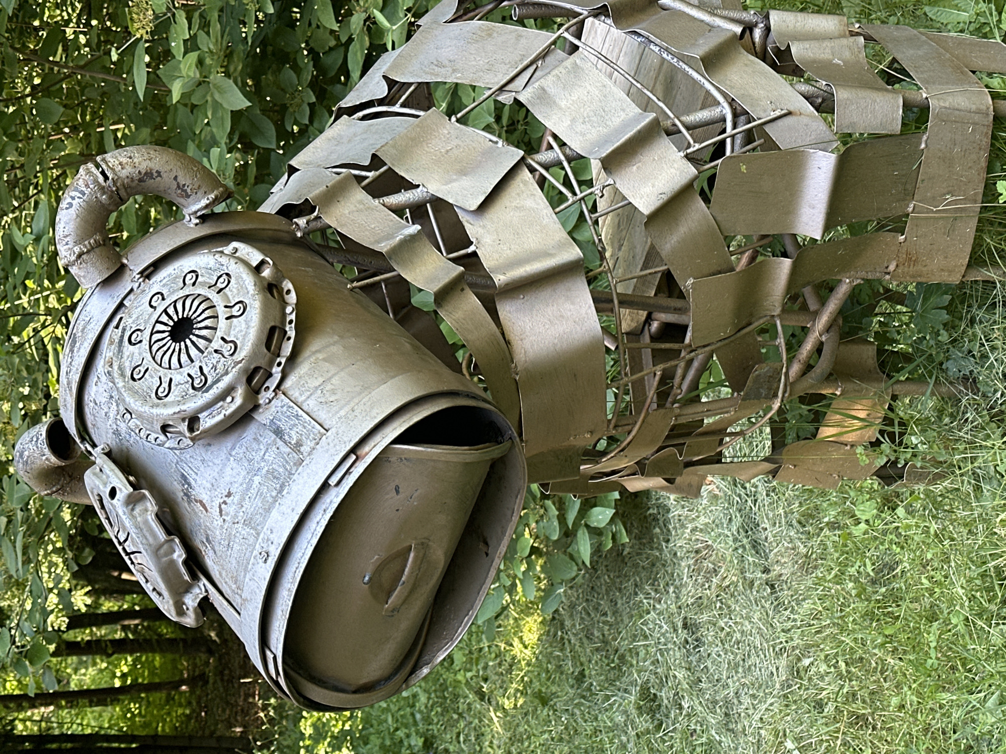 A metal sculpture of a dog is positioned outdoors in a grassy area with trees and foliage in the background. The sculpture is made from various recycled metal parts, including what appears to be a clutch pressure plate and a pipe forming the head, and a body constructed from overlapping metal strips. The dog is facing left, with its head tilted downwards. The environment suggests a park or garden setting.