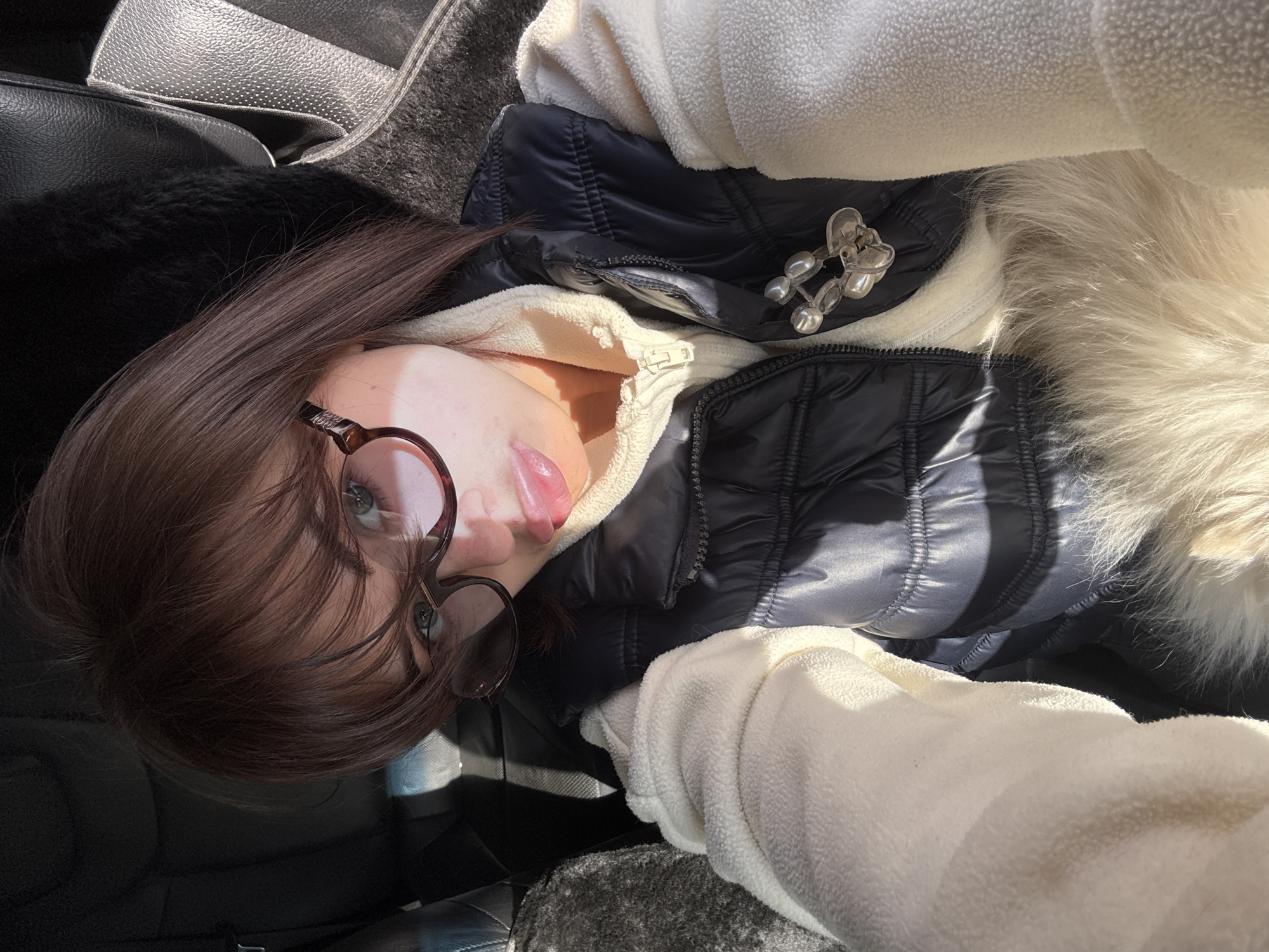A selfie-style photo taken indoors in Лобаново, Russia, shows a woman with brown hair and glasses lying down, partially visible in the frame. She's wearing a cream-colored fleece and a dark, quilted jacket with a decorative brooch. A fur-trimmed edge is also visible. To the left, parts of a black car seat are visible.