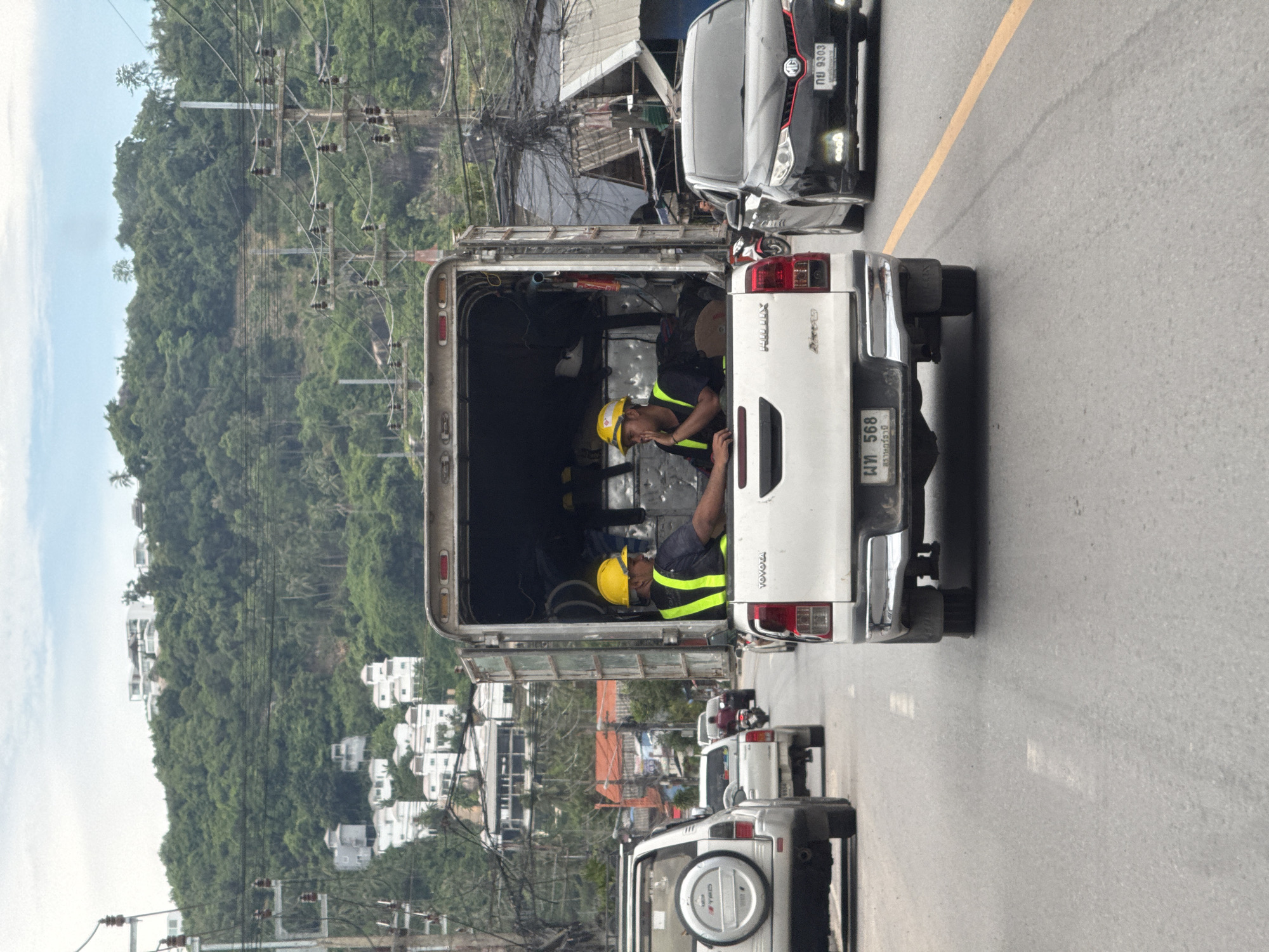 In Baan Hua Thanon, Thailand, two men wearing yellow hard hats and reflective vests are seated in the open bed of a white pickup truck on a paved road. The man on the left is looking forward and appears to be speaking, while the man on the right is looking to his left and resting his chin on his hand. The truck is in traffic, with a white SUV in front of it and a black sedan to its right. In the background, a lush green hillside is dotted with white buildings and intersected by power lines. The sky is overcast with some patches of blue.