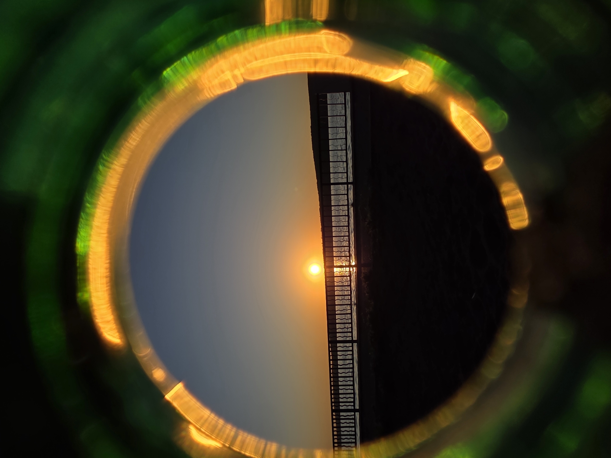 A sunset view is captured through a circular lens, showing a horizon line bisecting the scene. To the left of the horizon, the sky transitions from a bright orange near the sun to a muted blue, while on the right, a dark expanse suggests either land or a shadow. A structure reminiscent of a ladder or a fence runs vertically along the horizon line, appearing to bisect the sun's reflection on a body of water. The lens through which the image is taken is ringed with a diffuse yellow and green glow, creating a vignette effect. The scene is a view of sunset along the river in Chaikovsky, Russia.
