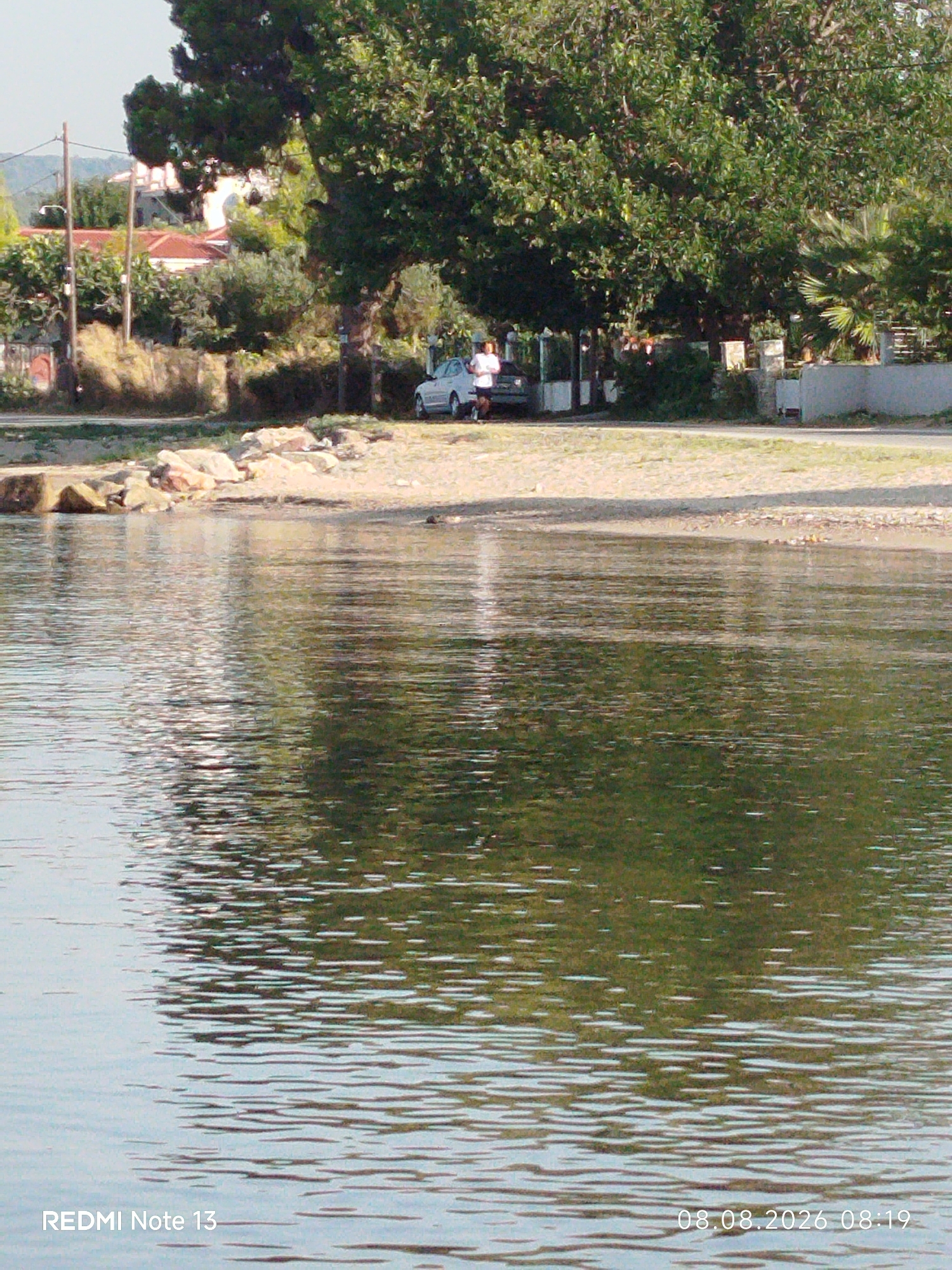 The image depicts a coastal scene, likely in Sithonia Municipal Unit, Greece, showcasing a body of water in the foreground with a textured surface reflecting the surrounding environment. In the background, a sandy shore is visible, transitioning to vegetation and buildings. A light-colored car is parked on the side of a road that runs parallel to the shore. A person, appearing to be a male, is standing next to the car. The overall impression is of a peaceful, outdoor setting during daylight hours.