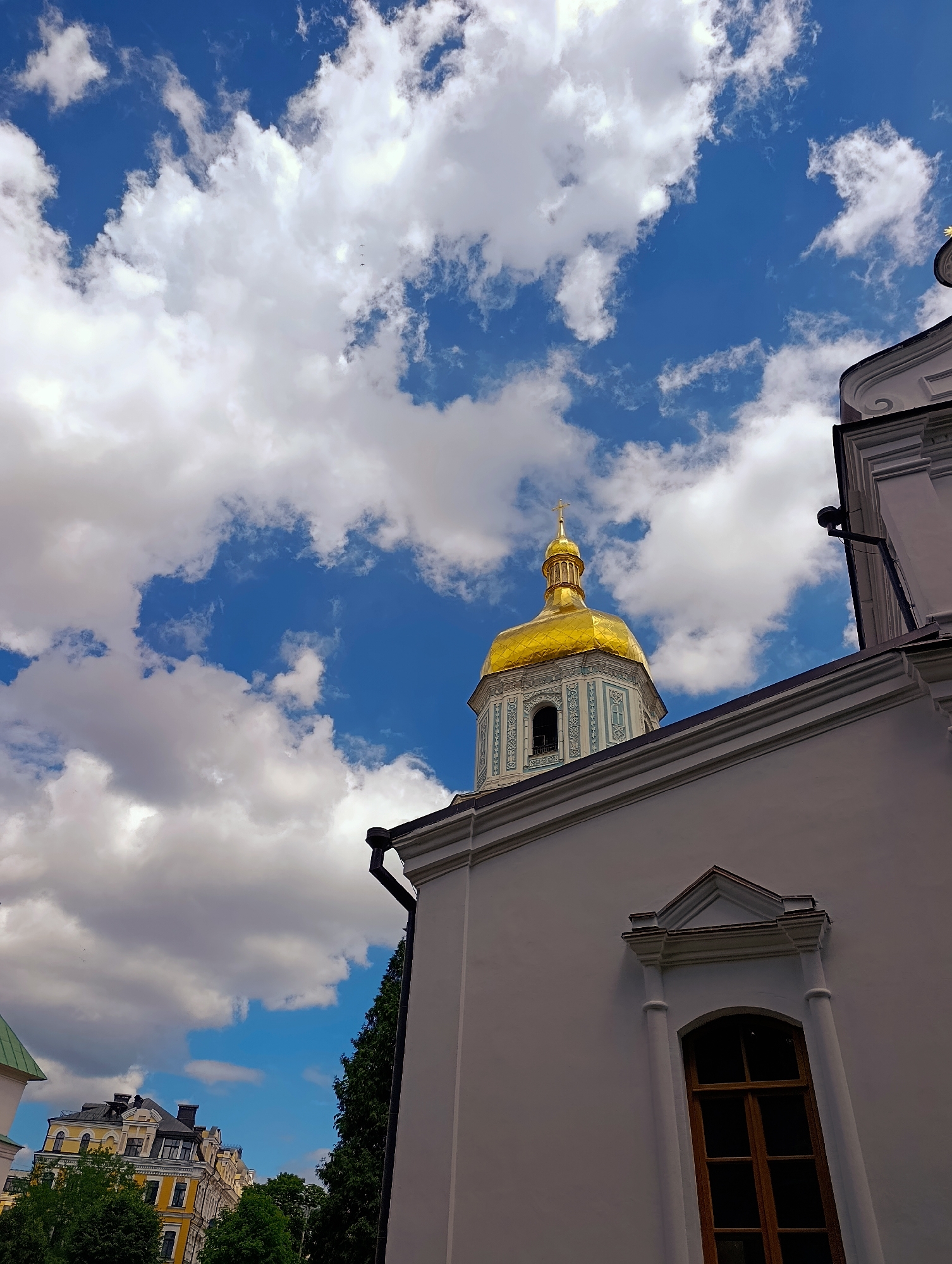This media file showcases a striking low-angle shot of St. Sophia's Cathedral in Kyiv, Ukraine, under a dynamic sky. The primary subjects are the cathedral's magnificent golden dome, reaching towards a vibrant blue sky dotted with fluffy white clouds, and the detailed architectural facade of the building. The scene is bathed in bright daylight, suggesting it was captured on a clear, sunny day, likely during the daytime given the illumination.
The golden dome, adorned with intricate carvings and topped with a cross, is the focal point. Adjacent to it is a section of the cathedral's white exterior wall, featuring an arched wooden window with a decorative pediment above it. The building's architecture is classical and ornate, hinting at its historical and religious significance. In the background, glimpses of other buildings with distinctive yellow facades and dark roofs can be seen, along with lush green trees, providing a sense of the urban setting.
The weather appears to be fair, with dramatic clouds adding visual interest to the sky. There are no people visible in the frame, focusing the viewer's attention on the grandeur of the cathedral and the sky. The overall mood is one of awe and tranquility, emphasizing the beauty and solemnity of the religious landmark. No visible text is present in the image.