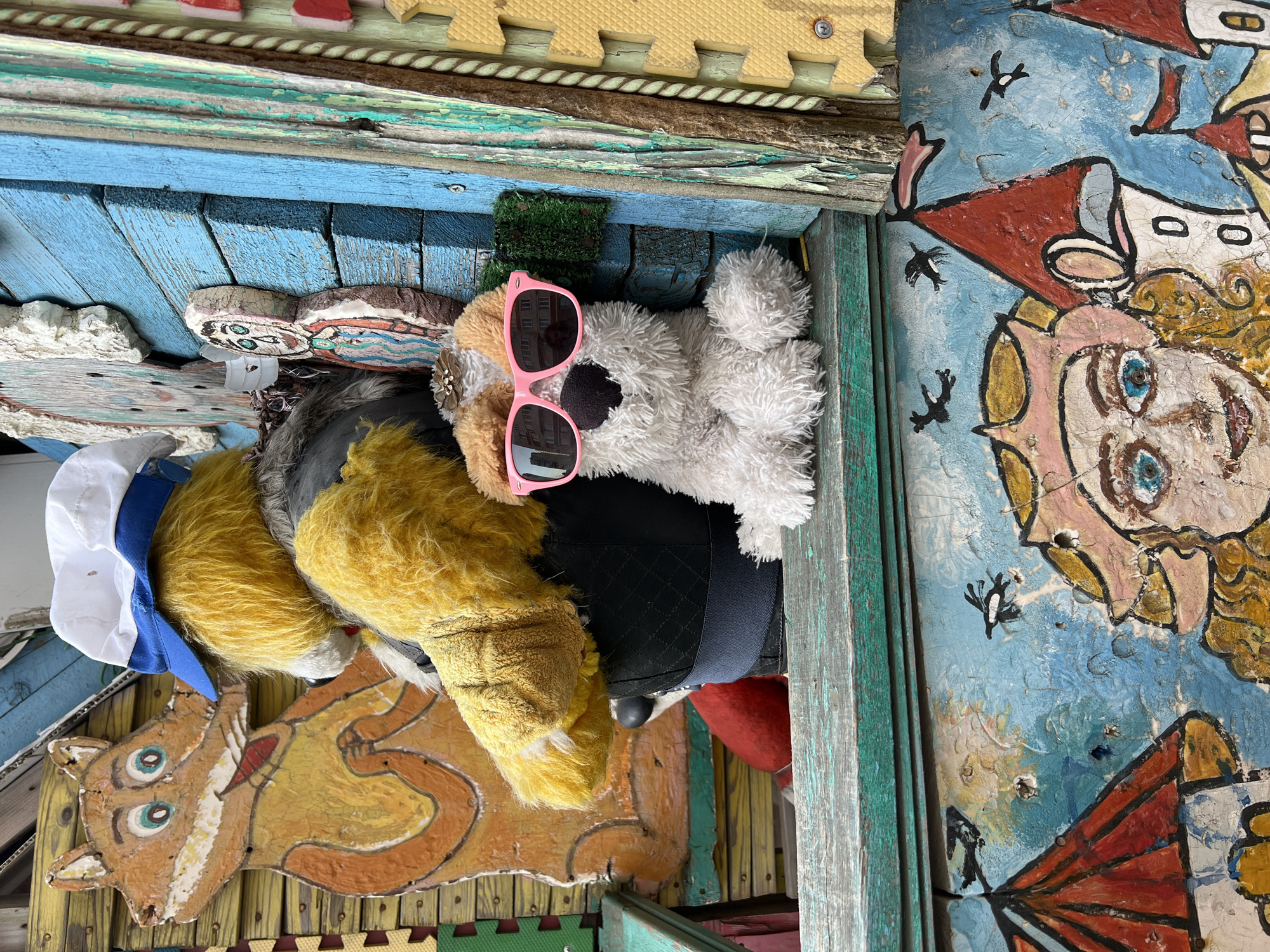 This image captures a vibrant and whimsical assemblage of objects, likely part of an outdoor art installation, a decorated playset, or a creative display. The original image is rotated 90 degrees counter-clockwise.
At the center of the arrangement lies a fluffy white stuffed animal, possibly a dog, wearing bright pink, oversized sunglasses. The lenses of the sunglasses clearly reflect an outdoor scene featuring multi-story buildings and a bright sky, suggesting the display is situated outdoors or near a window. Beside the white stuffed animal, to its left, another stuffed animal with shaggy yellow fur is visible, adorned with a white sailor's hat featuring a blue band. A small patch of green, moss-like material is nestled near the white stuffed animal's head.
The objects are set within a structure predominantly made of wood, which shows significant signs of wear, including peeling paint and weathered textures. The wooden surfaces are painted in various shades of blue and green, with some yellow hues also present, adding to the rustic and aged aesthetic. A textured, woven rope or trim runs along the upper edge of the wooden structure.
Several painted wooden cutouts are integrated into the scene. On the lower left, a stylized orange animal, resembling a fox or a cat, with prominent green-blue eyes and a white snout, is displayed. To the right, a brightly painted, human-like face, likely a girl with large blue eyes and curly blonde hair, is part of a larger painted backdrop. This backdrop depicts red-roofed buildings and several black bird silhouettes flying against a textured, light blue sky. Another smaller, more abstract painted figure is partially visible on the far left.
The entire display is framed by yellow foam puzzle mat pieces visible at the top and bottom edges. The materials and artistic style suggest a handmade, folk art quality, creating a cheerful and imaginative vignette. There is no visible text in the image.