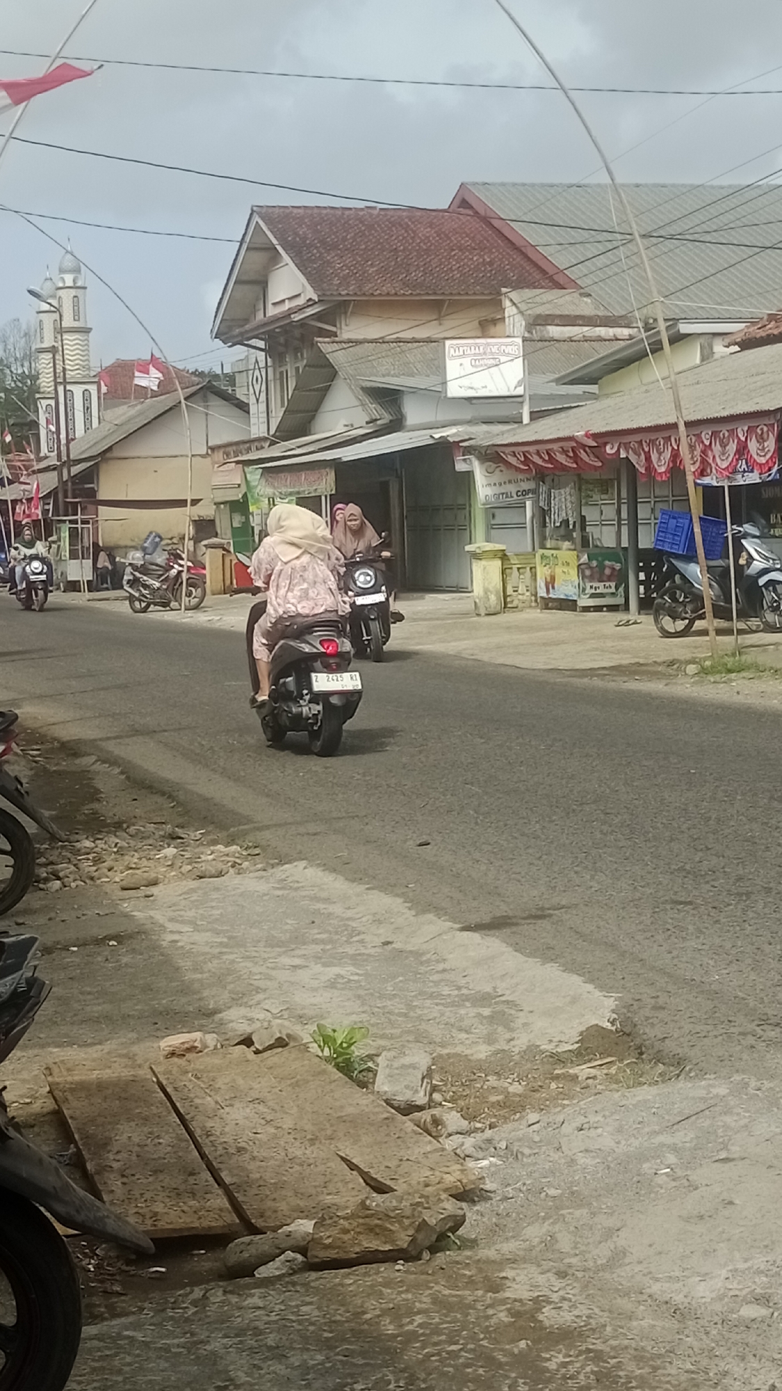 The image shows a street scene in Cikalong, Indonesia. In the foreground, two individuals are riding on a motorcycle. The person in the front is wearing a light-colored headscarf and a patterned shirt, while the person behind them is wearing a headscarf and a pink shirt. Both are facing away from the camera. The motorcycle has a license plate visible. To the left and right of the road, there are some parked motorcycles and buildings. One building has a sign that appears to be for a business. The street is paved and there are some signs of wear and tear. The overall scene suggests a typical day on a local street in Indonesia, with people going about their daily activities on motorcycles.