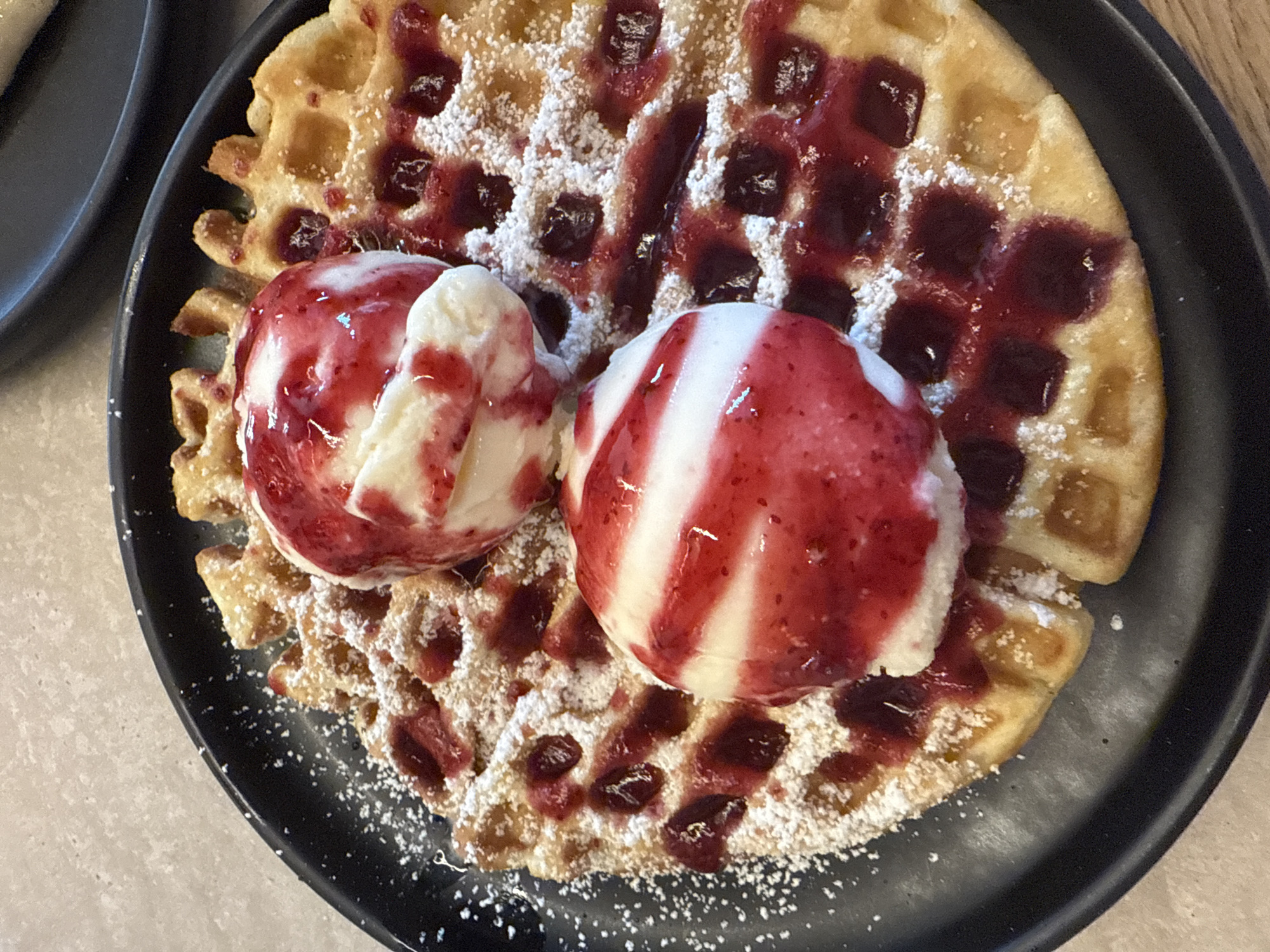 A close-up, overhead shot shows two scoops of ice cream with red syrup drizzled on top of a waffle dusted with powdered sugar and jam. The waffle is on a black plate, and the plate is on a light-colored, textured surface. The ice cream appears to be vanilla with swirls of red syrup. The jam on the waffle is dark red. The waffle has a grid pattern.