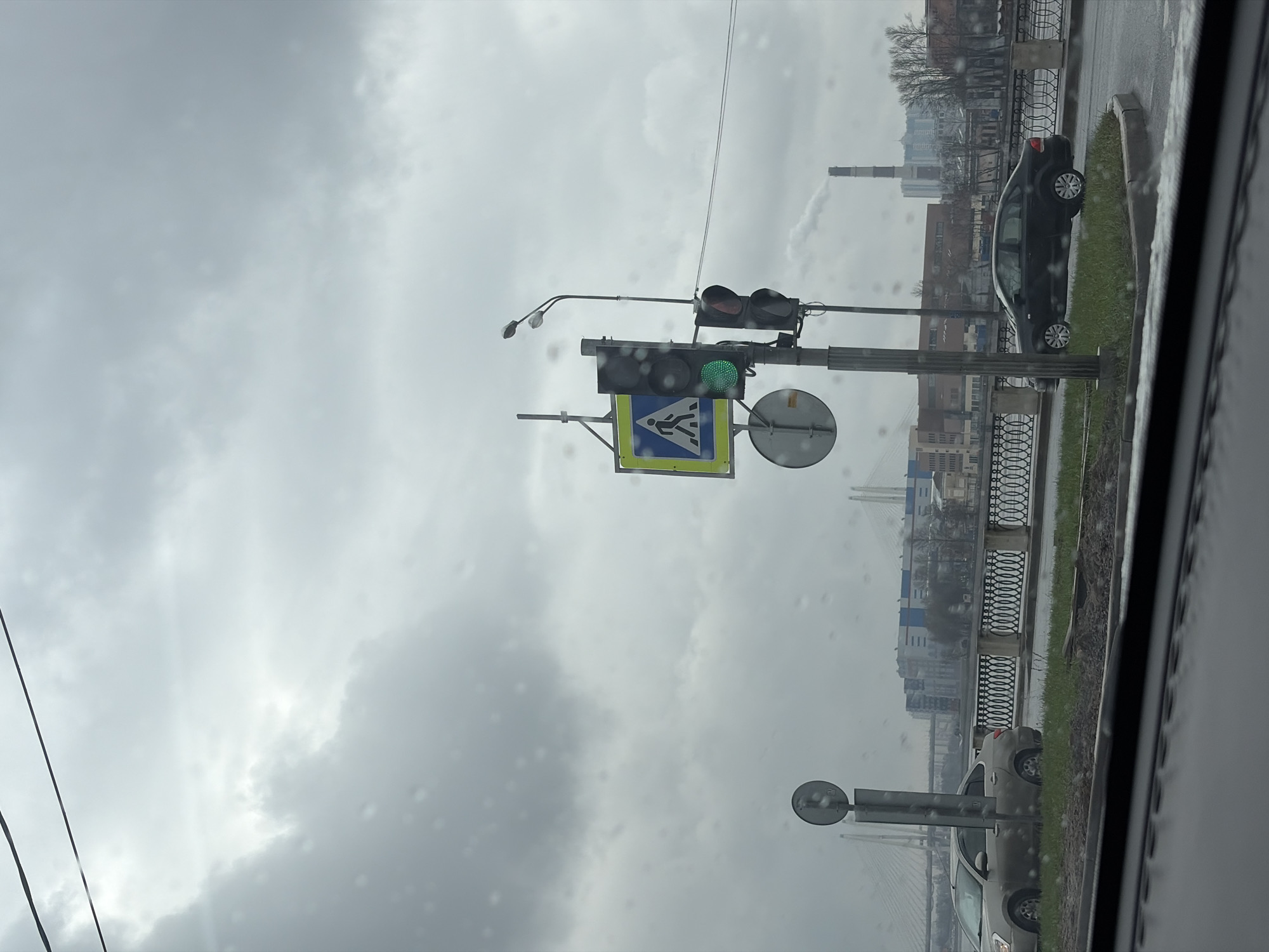 View from inside a vehicle on a gray, overcast day in Saint Petersburg, Russia. A traffic light shows green above a pedestrian crossing sign. Beyond the traffic light, a black car is parked on the grass next to a wrought iron fence bordering the embankment. In the background, a cityscape includes buildings and a smokestack, suggesting an industrial area. Rain is visible on the window. A bridge is seen in the far distance.