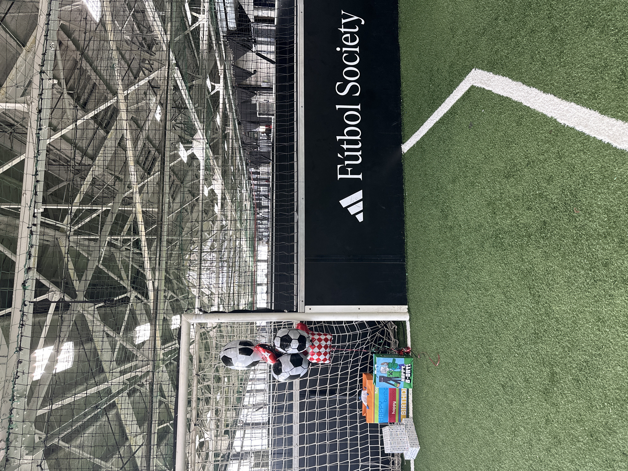 The image shows an indoor soccer field with a soccer goal in the foreground. The field is artificial turf with a white boundary line visible. A black banner with white text that reads "Fútbol Society" and an Adidas logo is mounted on the wall behind the goal. To the left of the goal, there are several birthday decorations, including balloons shaped like soccer balls and gift bags. The ceiling is industrial with exposed metal structures and lighting. No people are visible in the image. The location is not specified in the image, but the banner suggests it is a soccer facility.