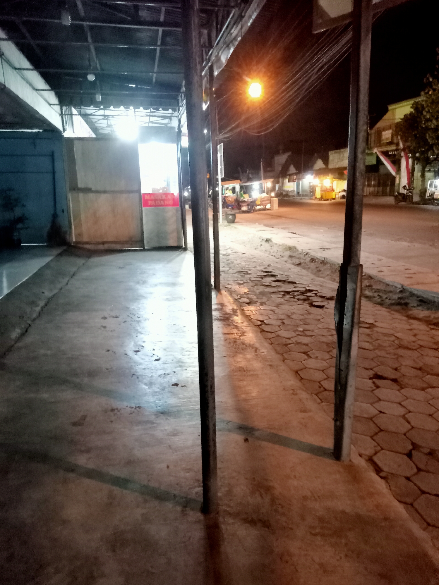 The image shows a nighttime view of a street in Mangunsari, Indonesia. The scene is illuminated by artificial lights from streetlamps and buildings. On the left, there is a covered walkway or awning with metallic supports. A blue wall is visible behind the walkway, and a red sign with white text that appears to read "MASAKAN PADANG" is mounted on a light-colored surface within the walkway. The ground in front of the walkway is a smooth, paved surface that appears to be concrete, with some scattered debris and dark spots.
To the right of the walkway, the street is paved with hexagonal interlocking blocks. A tall, dark metal pole stands prominently in the foreground, casting a shadow on the paved ground. In the distance, there are several buildings with lit windows, suggesting commercial establishments. A streetlamp with a bright orange glow is visible in the upper portion of the image, with overhead electrical wires crossing the sky. A small motorized vehicle, possibly a three-wheeled transport, is parked on the side of the road further down. Trees with sparse foliage are present on the right side of the street, and a flag with red and white stripes is visible on a building in the background. The overall impression is of a moderately busy commercial or residential street at night.