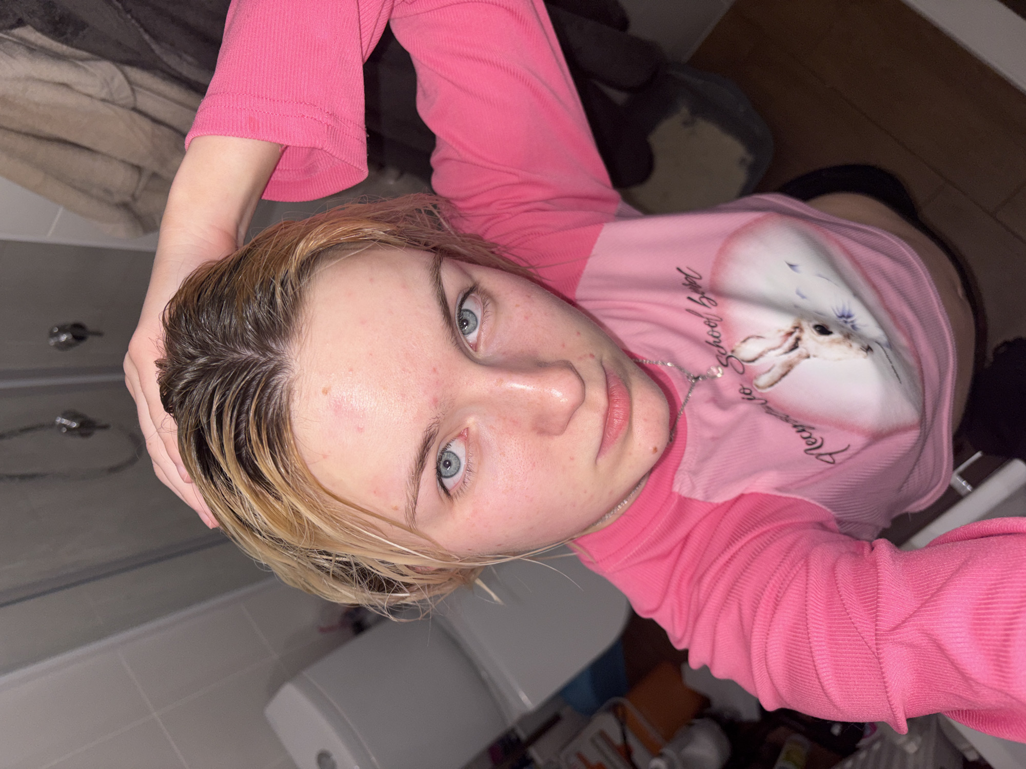 A high-angle, close-up selfie depicts a young person, likely female, with wet hair, dark at the roots and blonde towards the ends. She looks directly into the camera with striking blue eyes and a neutral expression. Her face shows some minor redness or blemishes.
She is wearing a ribbed, long-sleeved pink top. The top features a graphic design on the chest, including a heart shape with an animal (possibly a fawn or deer) inside, and partially visible text that appears to read "My School Beauty." A thin silver chain necklace is visible around her neck. Her left hand is raised, possibly holding or pushing back her wet hair.
The background suggests an indoor setting, likely a bathroom, with light-colored tiled walls and hints of fixtures such as a faucet or shower head on the left and a toilet or sink on the right. The lighting is artificial. The image captures a moment, possibly after washing her hair. The location context provided is Ligolambi, Russia.