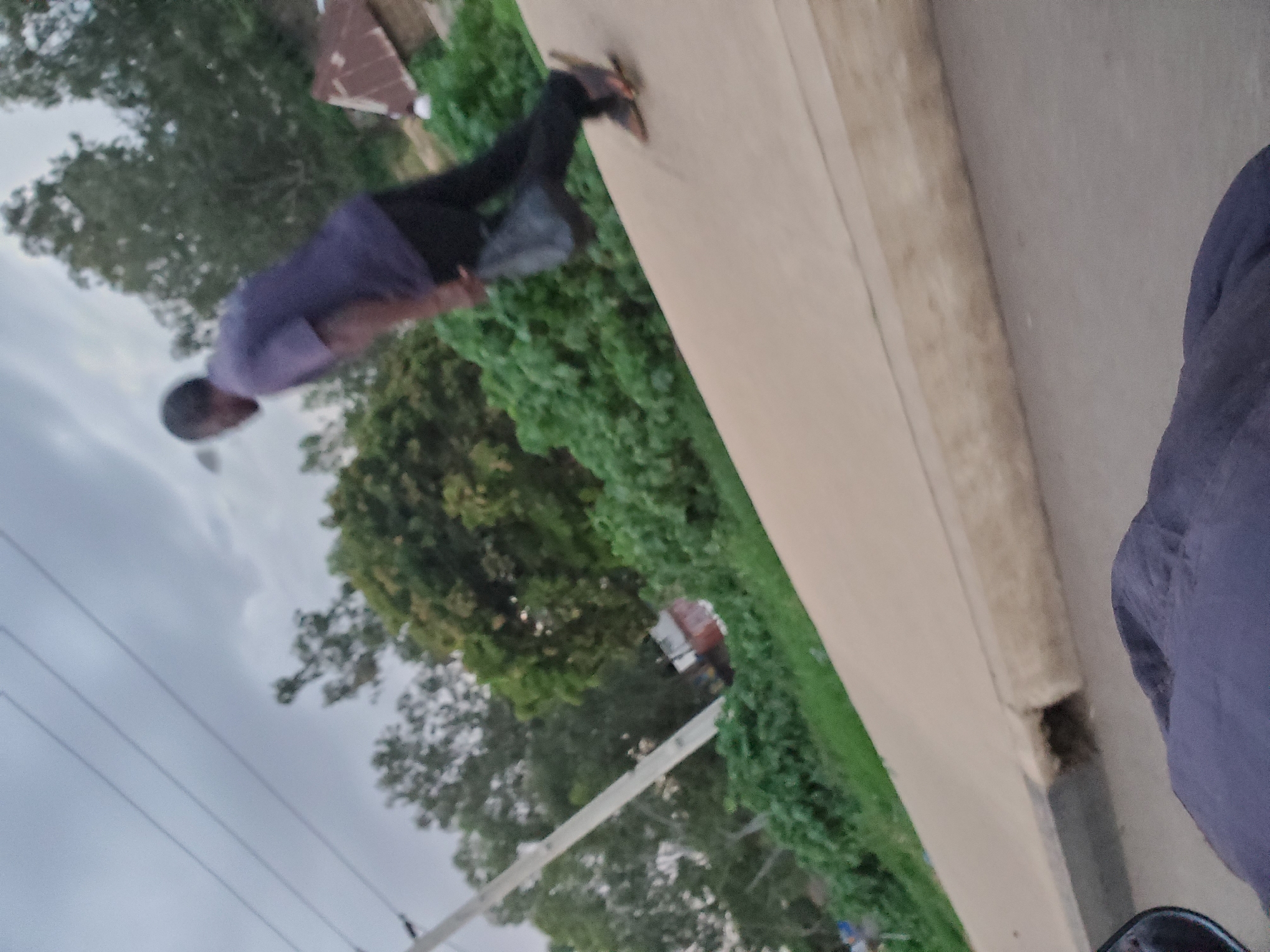 The media file shows a person performing a handstand against a wall. The subject is wearing a purple shirt and black pants, with dark shoes. The person appears to be outdoors, with greenery and trees visible in the background. There is also a glimpse of a building with a corrugated iron roof, and utility lines crossing the sky. The sky is overcast, suggesting it might be a cloudy day, possibly in the late afternoon or early morning. The overall scene appears to be in a residential or semi-rural area. The person's posture and the fact that they are in a handstand suggest they are engaged in physical activity, possibly exercise or a stunt. There is no visible text in the image. The location context is Gura Top, Nigeria.