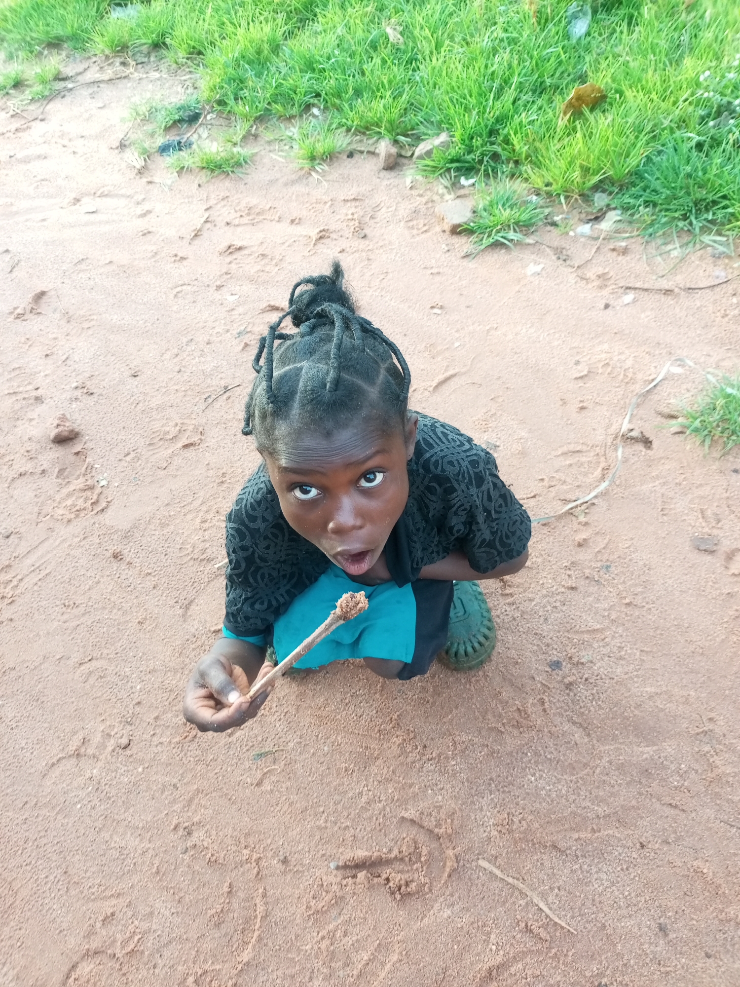 A young child is crouched on reddish-brown earth, looking upwards at the camera with a surprised expression. The child has dark skin, braided hair, and is wearing a black patterned top over a turquoise garment, and green footwear. They are holding a twig with some material stuck to its tip. The ground appears to be a mixture of sand and small pebbles, with patches of green grass visible in the upper portion of the image. This scene is located on a dirt path in Tempure, Nigeria.