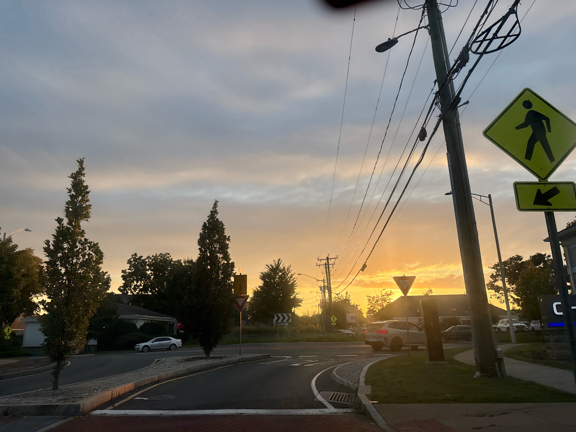 A street-level, slightly wide-angle photograph captures a sunset over a suburban intersection in Glastonbury, Connecticut, United States of America. The sky is a blend of muted grays and soft oranges, with wisps of clouds illuminated by the setting sun. Utility poles and their associated wires dominate the right side of the frame, stretching across the sky. A prominent diamond-shaped yellow pedestrian crossing sign with a black silhouette of a person walking is visible on the right, accompanied by a smaller rectangular sign below it displaying a black arrow pointing left.
In the foreground, a paved road curves through the intersection, marked with white lines. To the left, a sidewalk with brick and stone edging separates the road from a grassy area. Several trees, including two tall, slender evergreen trees, line the roadside. Behind the trees on the left, residential houses with visible roofs are partially obscured by foliage.
Several vehicles are present. A silver sedan is parked on the left side of the road. Further down the street, a white SUV is turning at the intersection. Another dark-colored SUV and a white truck are visible in the background. A circular yield sign is positioned at the intersection. On the right side of the frame, a portion of a building with illuminated blue lettering that appears to read "C-" is visible. The overall scene depicts a typical evening transition in a suburban environment, with the warm glow of the sunset contrasting with the utility infrastructure.
