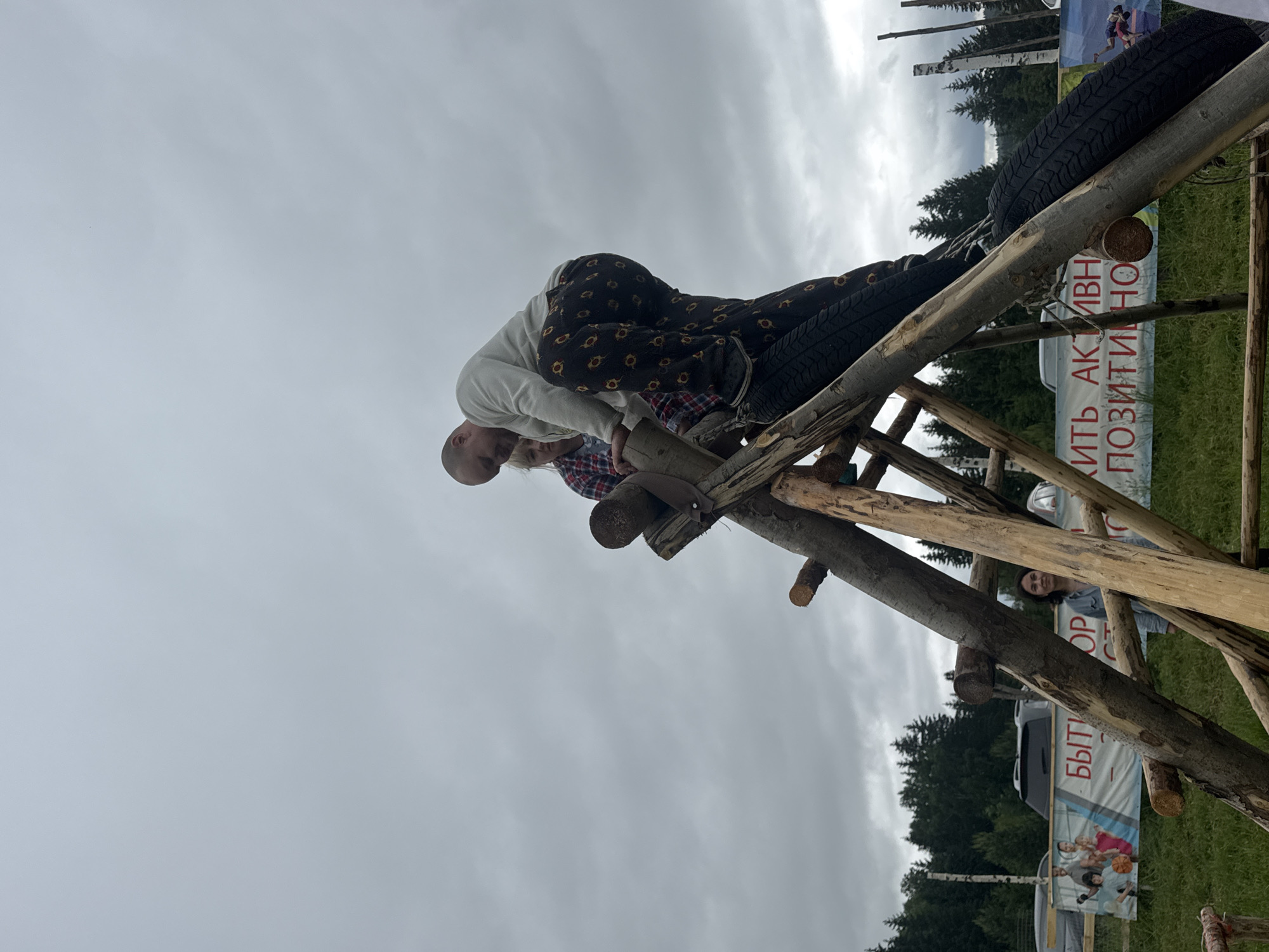The image depicts an outdoor scene in Большая Кивара, Russia, under an overcast sky. In the foreground and mid-ground, a handcrafted wooden climbing structure, made from rough-hewn logs, stands centrally. Integrated into the structure are black car tires, serving as climbing elements.
On the upper part of the wooden structure, a bald man wearing a light-colored long-sleeved shirt and dark pants with a yellow circular pattern is actively climbing or standing, with his left foot on one of the tires. Behind and slightly below him, a woman with light hair, wearing a red and black plaid shirt, is also positioned on the structure.
In the mid-ground to the left, standing on green grass behind the structure, another woman with dark hair, wearing a light-colored long-sleeved shirt, looks directly towards the viewer with a slight smile.
In the background, two large banners are visible. The left banner shows illustrations of people, including children playing basketball, and partial Russian text "БЫТЬ" and "ОР". The right banner displays partial Russian text "ИТЬ АКТИВНО" and "ПОЗИТИВНО", which translates to "BE ACTIVE" and "POSITIVELY". Behind the banners, several vehicles, including a grey SUV, are parked. A dense line of coniferous trees forms the distant background, with scattered wooden poles visible throughout the scene. The ground is covered in green grass, consistent with an outdoor park or recreational area.