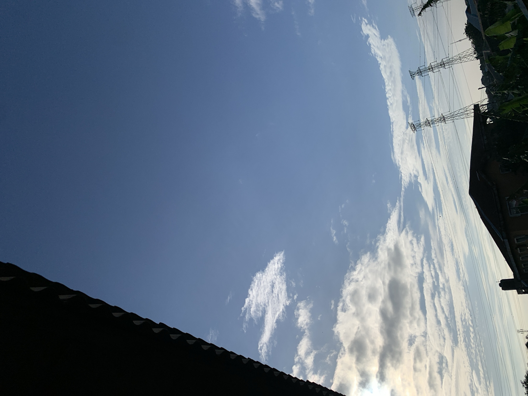 A sideways view of Ikorodu, Nigeria, reveals a bright, mostly blue sky interspersed with white clouds. A section of dark, tiled roof occupies the lower left corner, contrasting with the light sky. Power lines and towers extend from the upper right edge of the frame, near a building and trees.