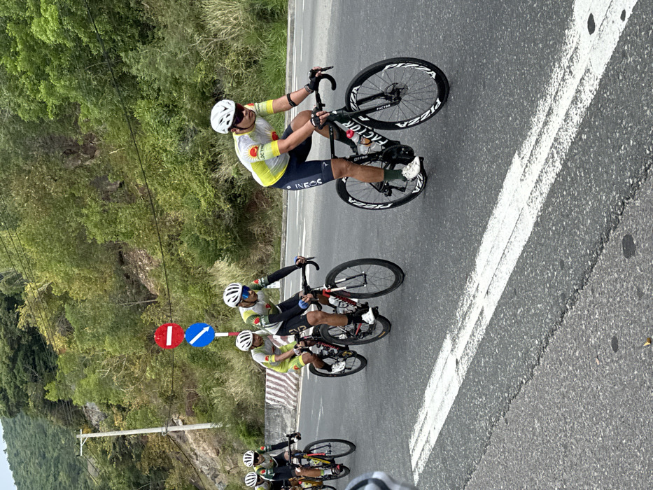 The image displays five cyclists in various stages of a group ride on an asphalt road, set against a mountainous, vegetated backdrop in Unknown, Vietnam. The foreground shows a partial view of a sixth cyclist's wheel and leg on the far right. The lead visible cyclist, positioned prominently on the right, wears a yellow, white, and red short-sleeved jersey, dark blue shorts with "INEOS" branding, a white helmet, and dark sunglasses; he rides a black "FACTOR" brand road bicycle. Behind him, four additional cyclists follow in a staggered single-file formation. They wear similar yellow, white, and red jerseys, with some featuring a red sun logo and "LOC TRI" text, along with white helmets and sunglasses. All are actively pedaling their road bicycles. To the left of the road, two circular traffic signs are mounted on a pole: a red "Do Not Enter" sign above a blue "Mandatory Pass on this Side" sign with a white arrow. A concrete barrier with red and white stripes is visible behind the signs, at the base of a steep, densely vegetated hillside. The road features double solid white lines marking a lane boundary. The sky is overcast.