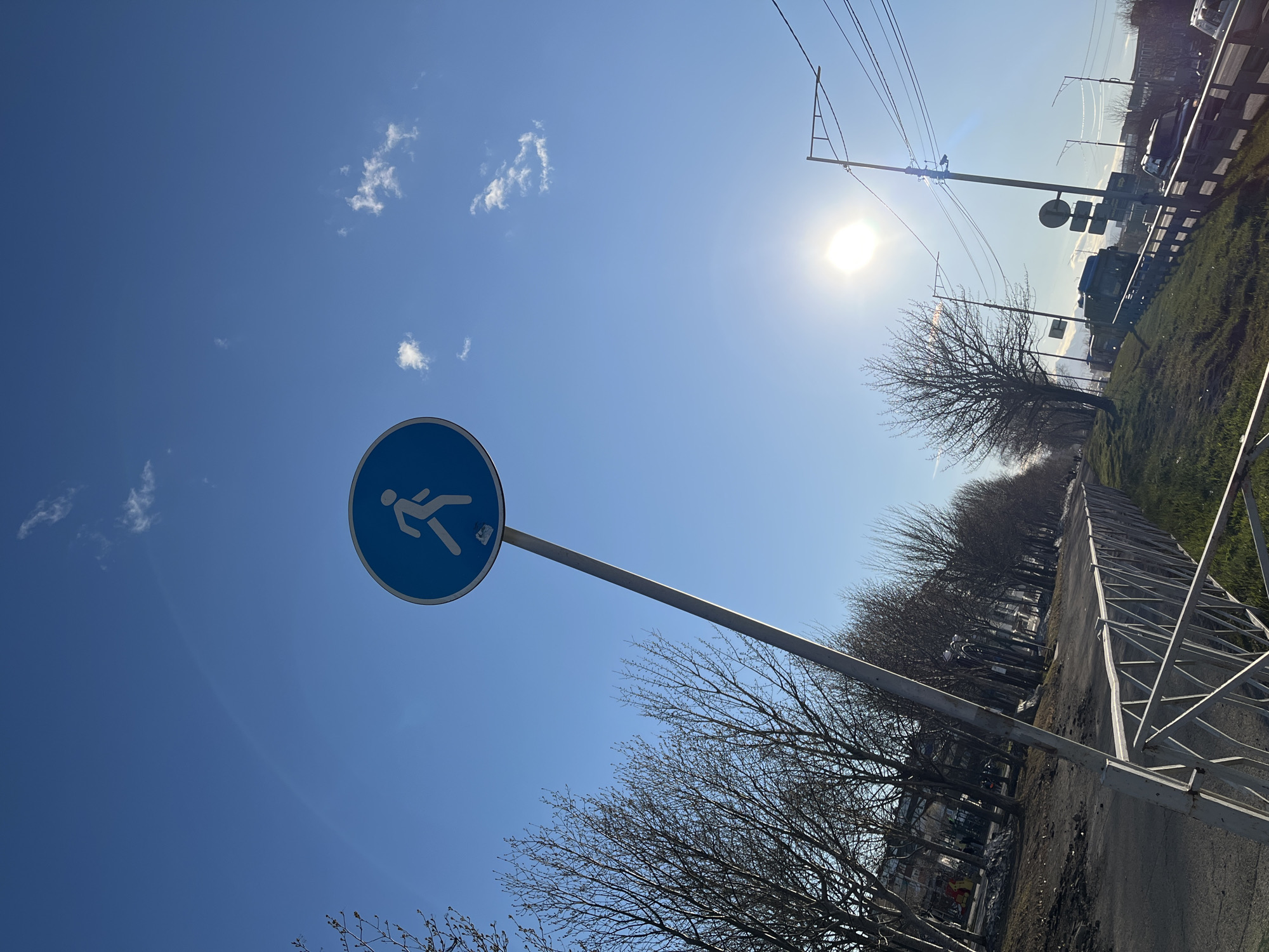 The image depicts a bright, sunny day in Petropavlovsk-Kamchatsky, Russia. In the foreground, there is a blue circular sign indicating a pedestrian zone, mounted on a silver pole. In the background, there are trees without leaves lining the road. In the upper right corner, there is an electric pole and some buildings in the far distance. The sky is a clear blue with a few wispy clouds. The sun is high in the sky, creating a bright glare. The shot appears to be taken from a low angle, emphasizing the height of the sign and the open expanse of the sky. The scene appears to be along a sidewalk or path adjacent to a road.