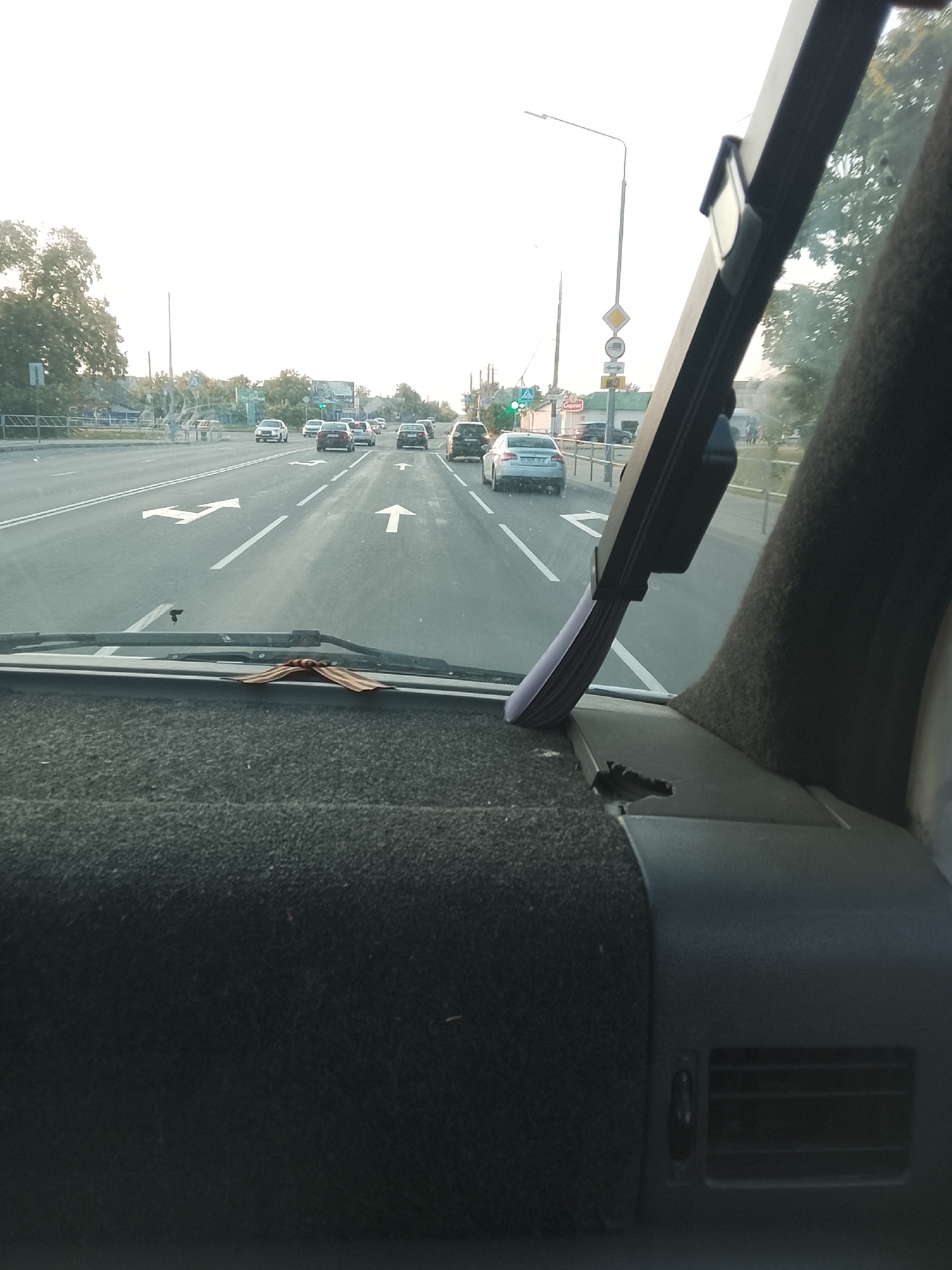 This is a point-of-view shot from inside a vehicle, looking forward through the windshield at a road in Zhlobin, Belarus. The road has multiple lanes and is marked with directional arrows. Several cars are visible on the road, traveling in the same direction. A traffic light is visible in the distance, which appears to be green. On the right side of the road, trees and buildings can be seen. The sky is bright, suggesting it is daytime, possibly late afternoon due to the soft light. The dashboard of the vehicle is visible in the foreground, covered in dark fabric. A decorative element, resembling a bow or ribbon, is placed on the dashboard. The overall scene depicts a normal driving situation on a highway. There is no discernible text visible in the image.
