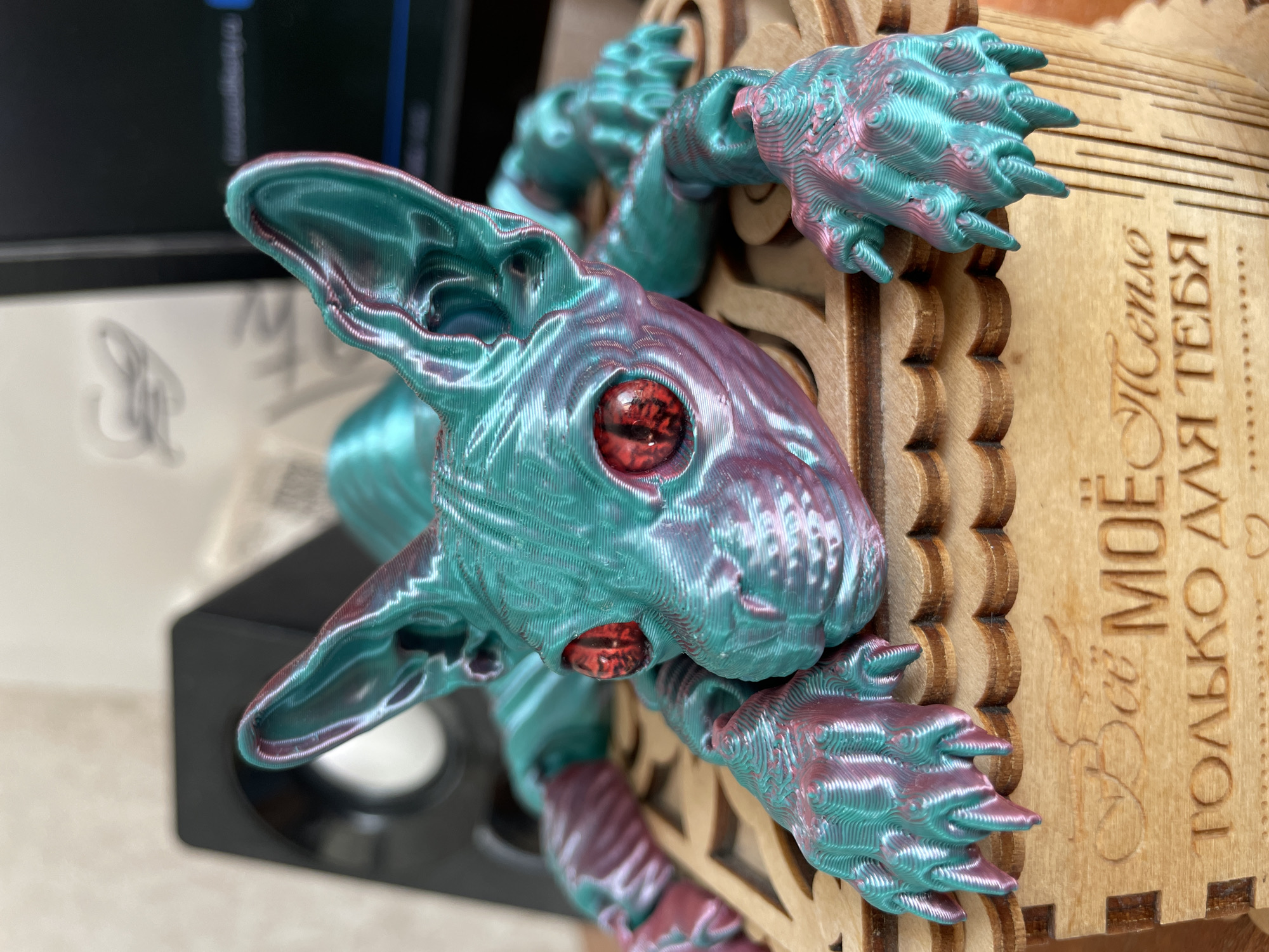 A close-up shot showcases a 3D-printed figure resembling a fantastical creature, possibly a stylized cat or rabbit-like form with elongated ears. The figure is predominantly colored in a turquoise-like hue with accents of pink or purple that appear to shift based on the light. Its eyes are strikingly red. The creature appears to be clinging to the side of a wooden box or structure, which has ornate cut-out designs. The box also bears Russian text, "Всё моё только для тебя," which translates to "All of mine only for you." The photograph is taken indoors, with a blurry computer monitor visible in the background. There is no location of Perm, Russia, identified in this image.